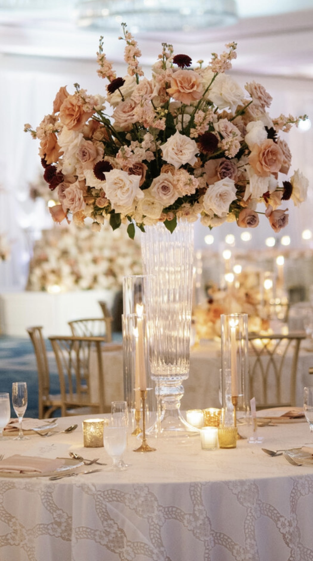 Tall floral centerpiece with pink and white flowers on a decorated table, candles, and elegant chairs in background.