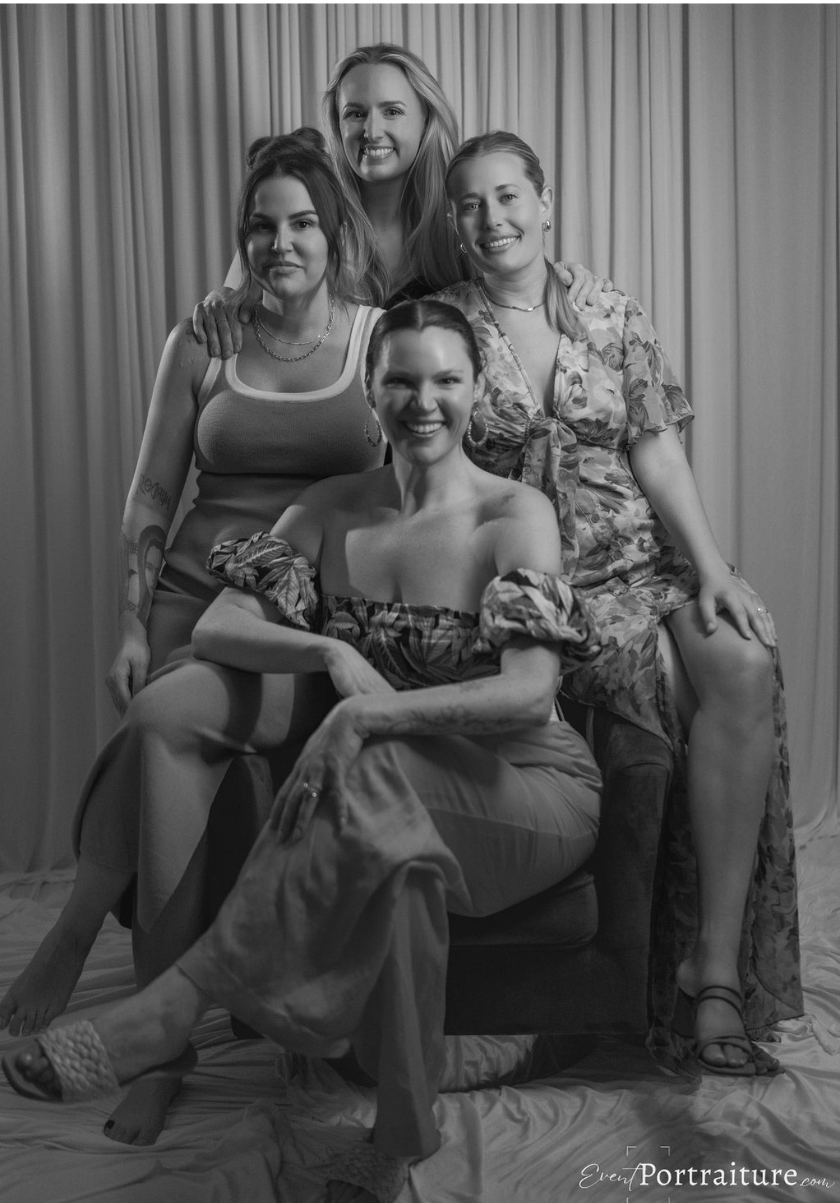 Four women pose together in front of a curtain, smiling, with one seated and three standing behind her.