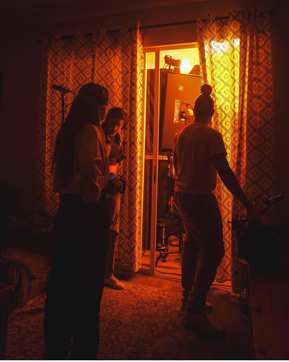 Four people stand in a dimly lit room with orange light coming from an open door, curtains, and furniture visible.