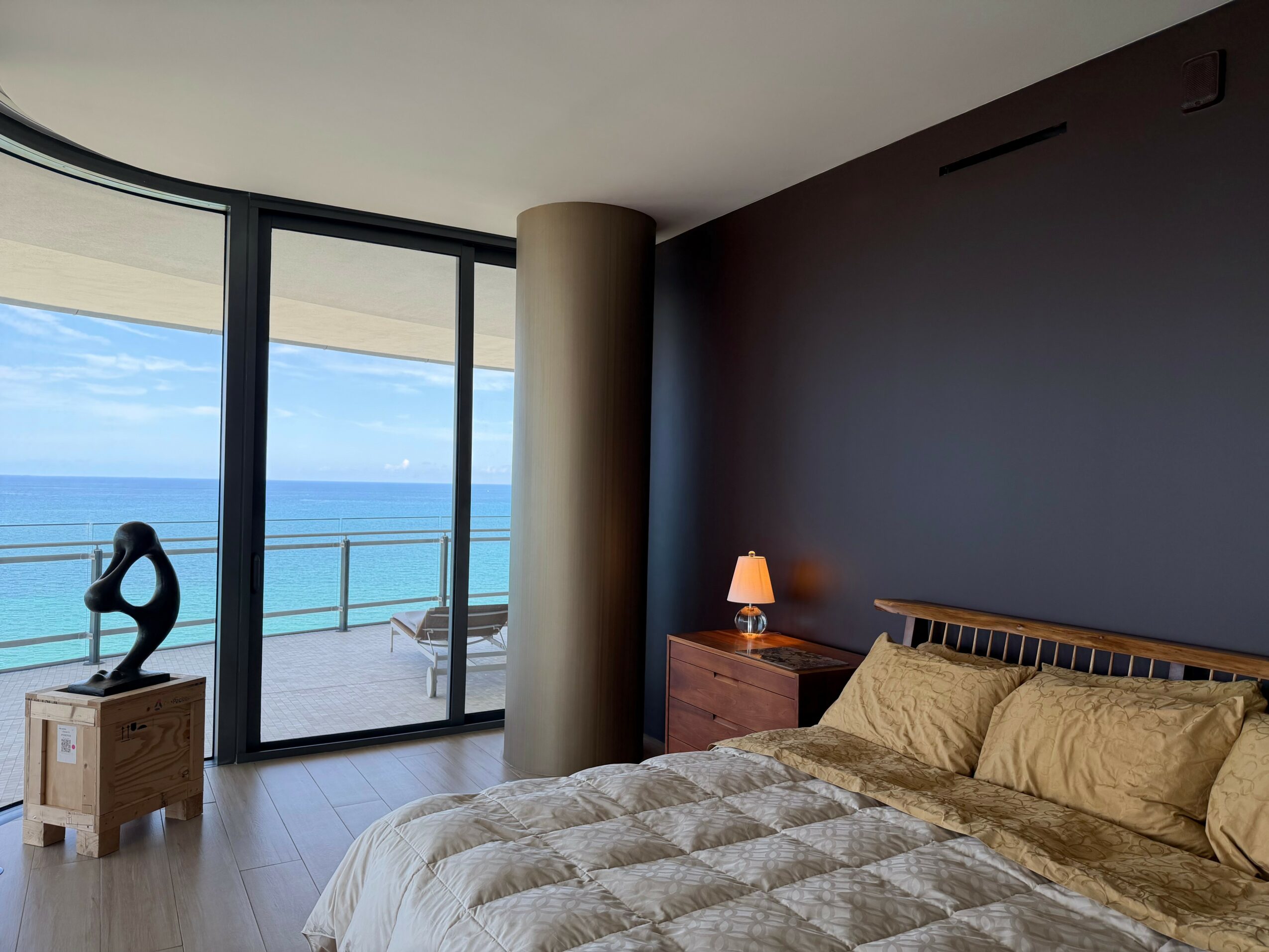 Bedroom with large glass doors opening to a balcony overlooking the ocean, bed with beige bedding, nightstand with lamp, and dark wall.