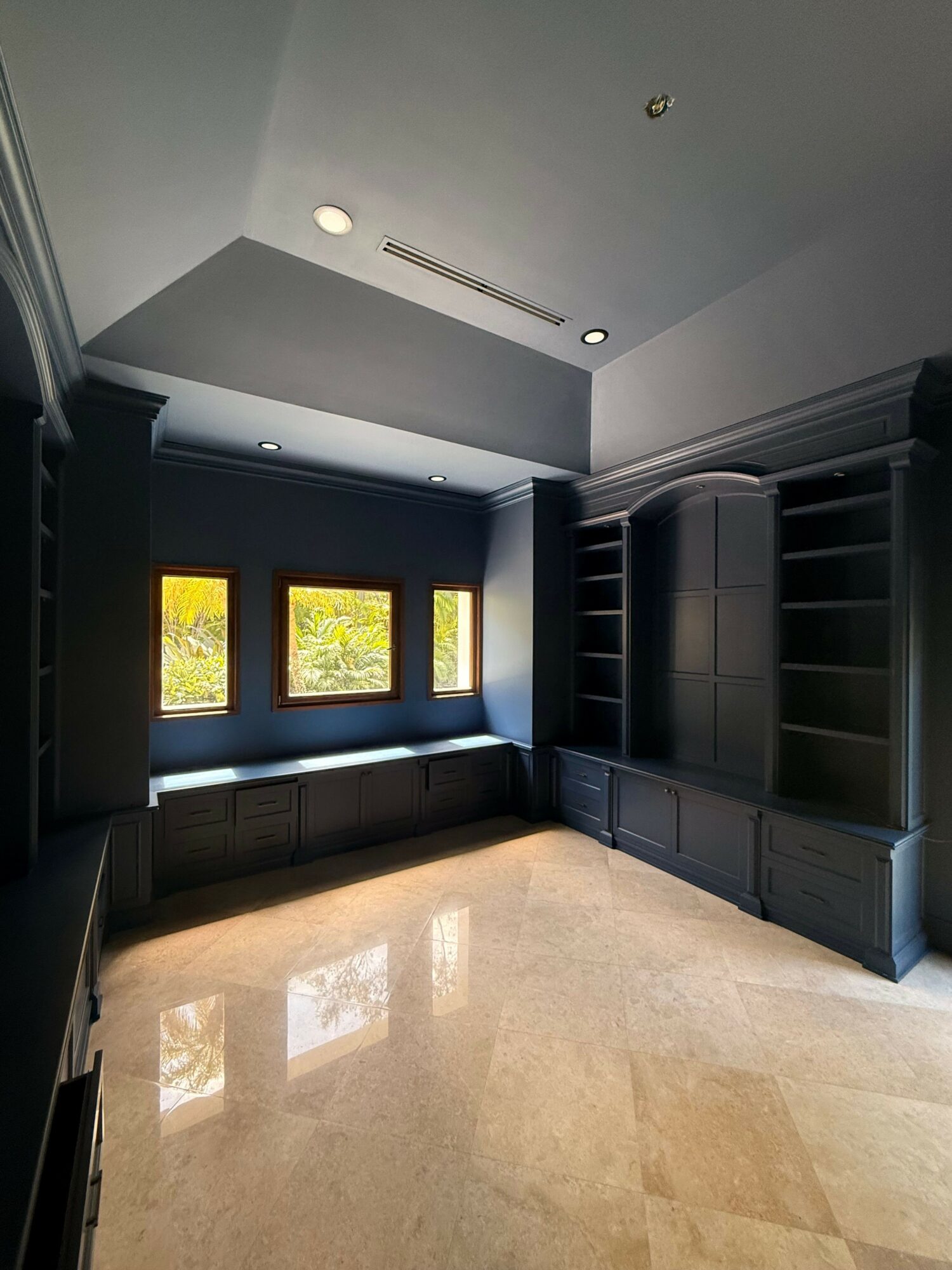 Room with gray walls, large gray cabinet, three windows showing trees outside, and a beige marble floor.
