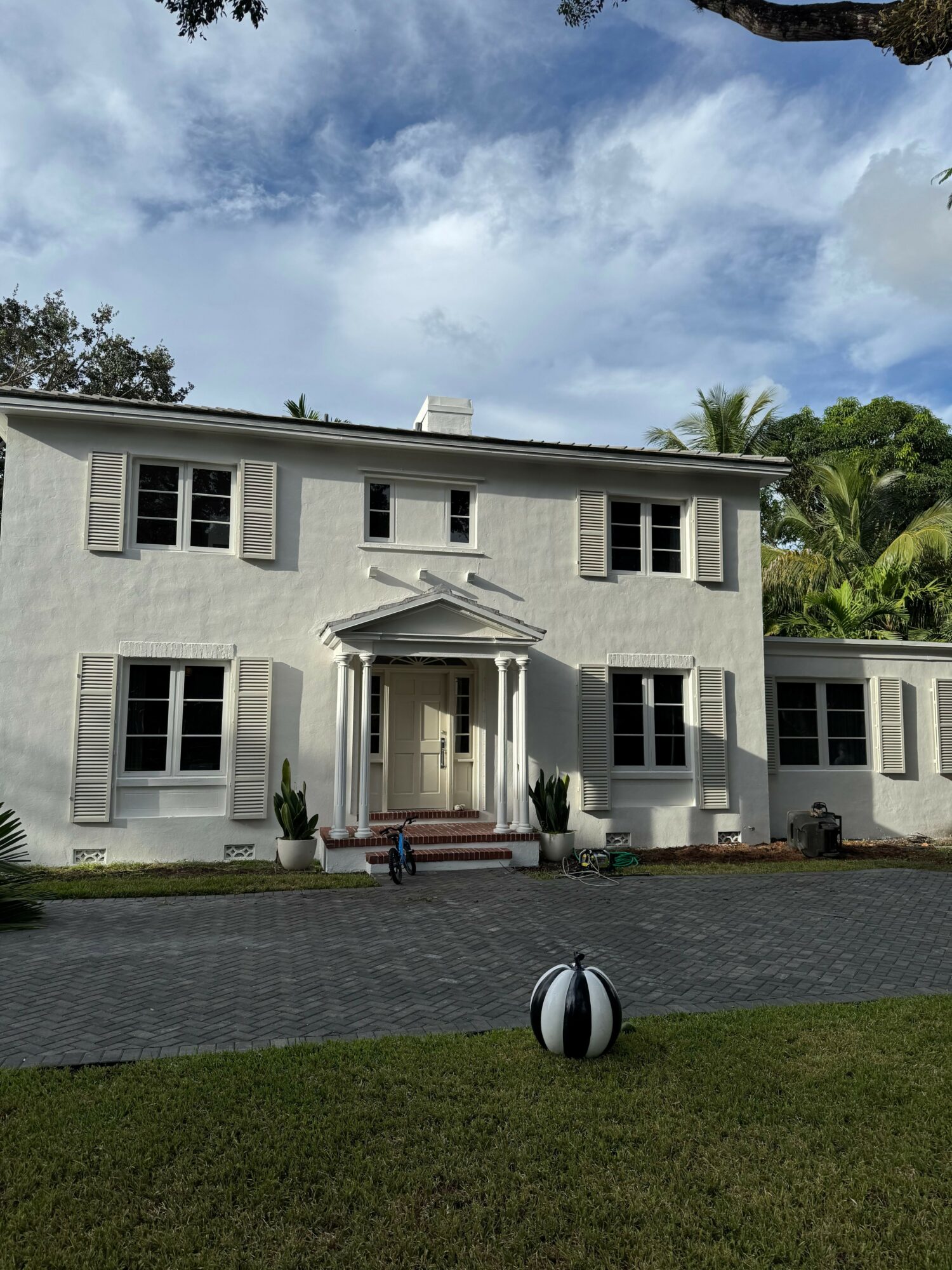 White two-story house with a front porch, surrounded by a lawn and trees, under a partly cloudy sky.