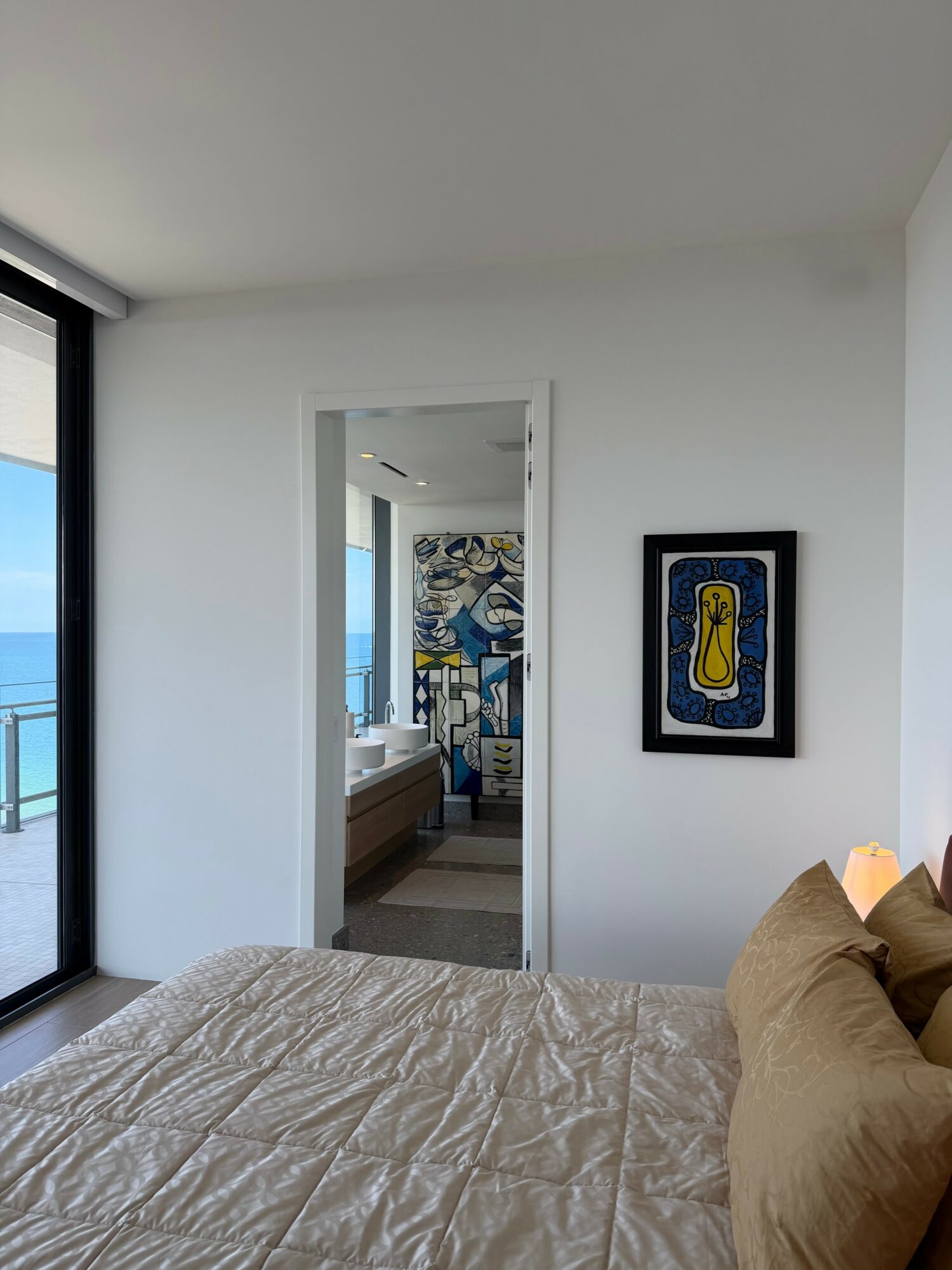 Bedroom with a bed, pillows, and a framed artwork on the wall, with a view of the ocean through a sliding glass door.