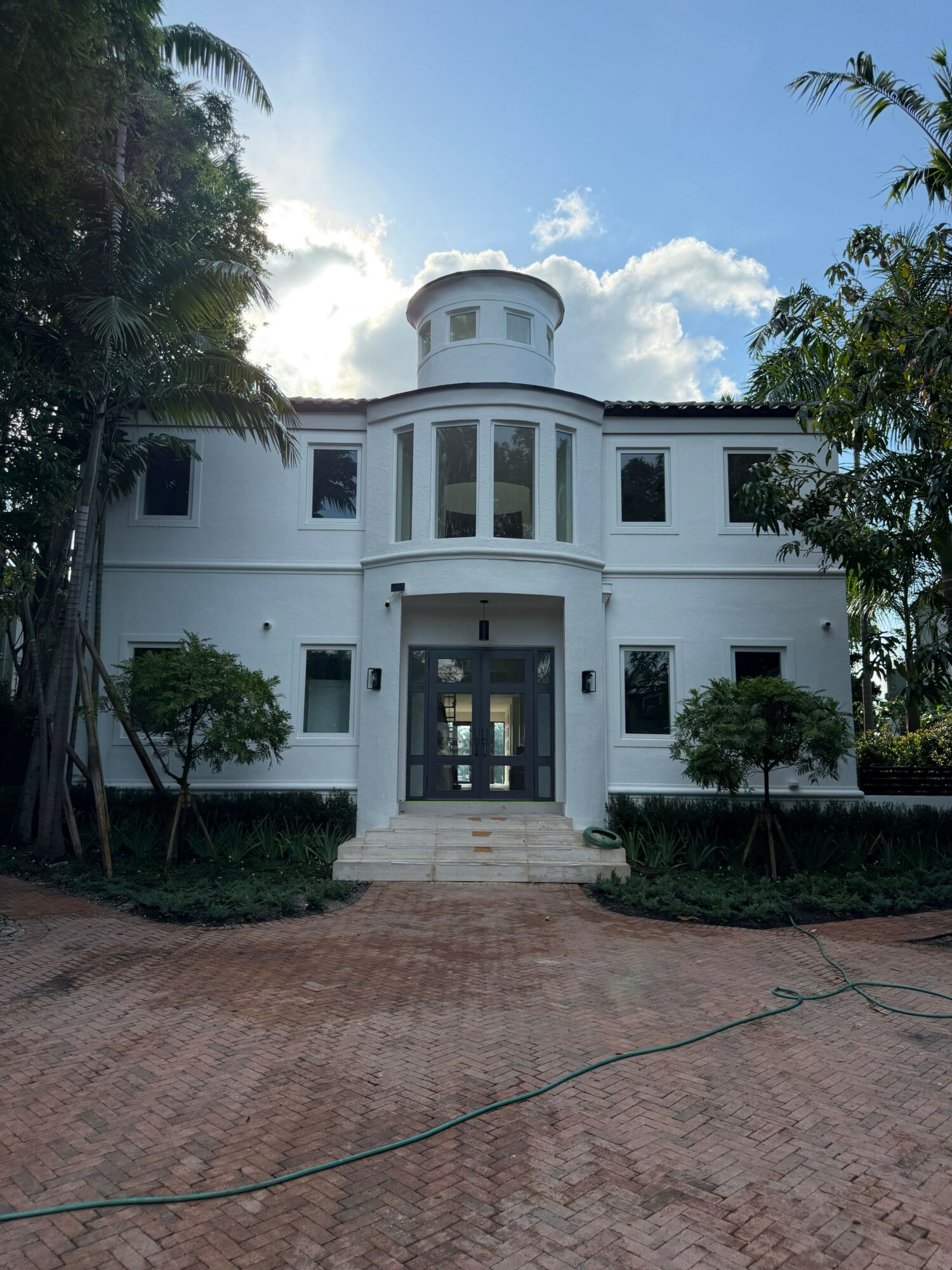 White two-story house with a rounded tower, black front door, and small trees in front yard, brick driveway, blue sky.