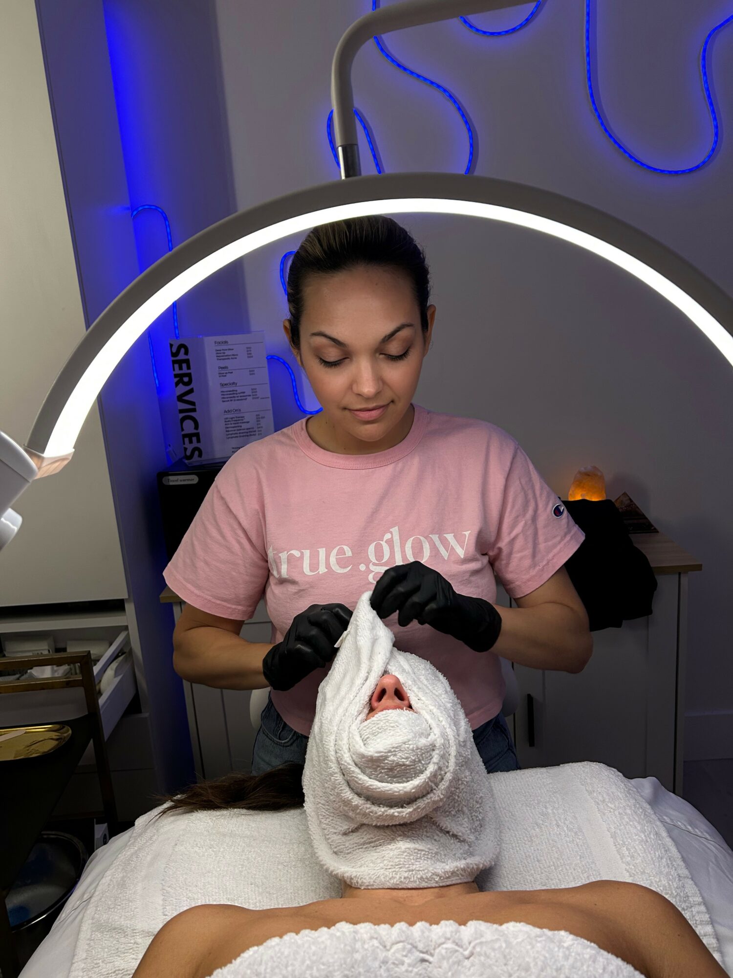 A person lying down with a towel wrapped around their head, receiving a facial treatment from a technician in a spa setting.
