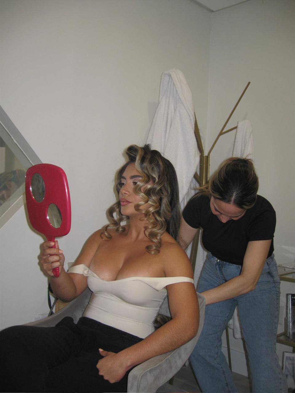 Woman with curly hair looking in mirror, sitting, with another woman adjusting her outfit in a room.