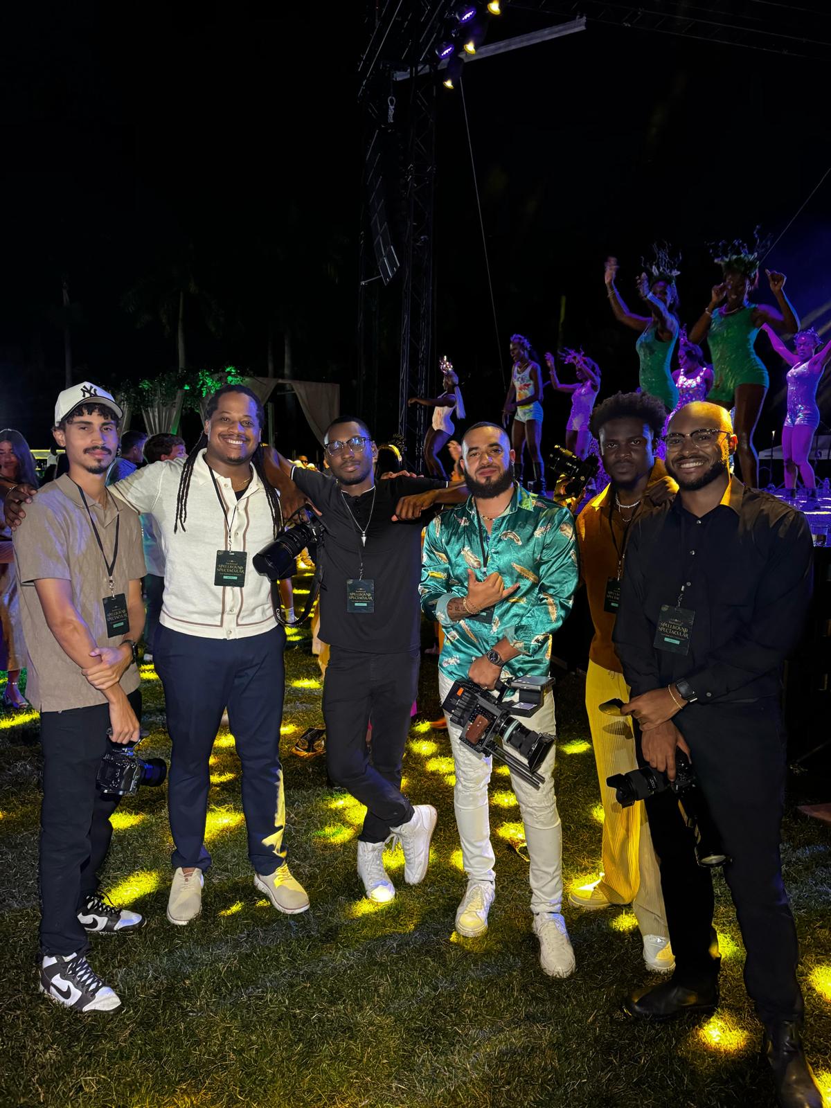 Group of six people standing outdoors at night, some with raised hands, with performers in colorful costumes in the background.
