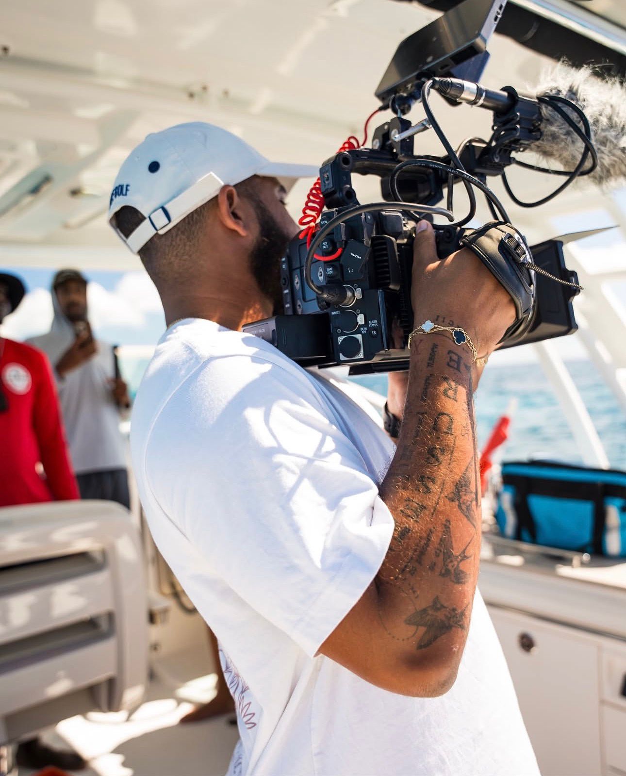 Person wearing a white helmet and shirt operating a professional video camera on a boat, with people in the background.
