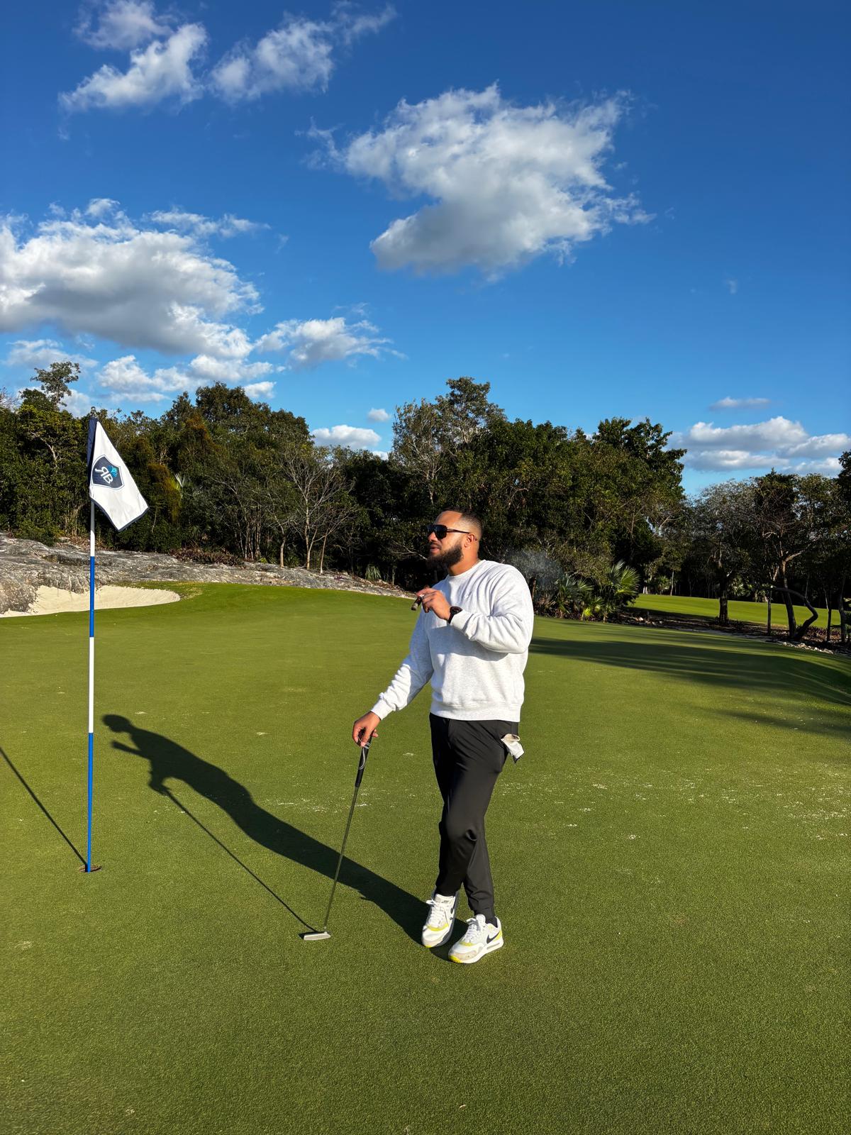 Person standing on a golf course near a flag, holding a golf club, with trees and blue sky in background.