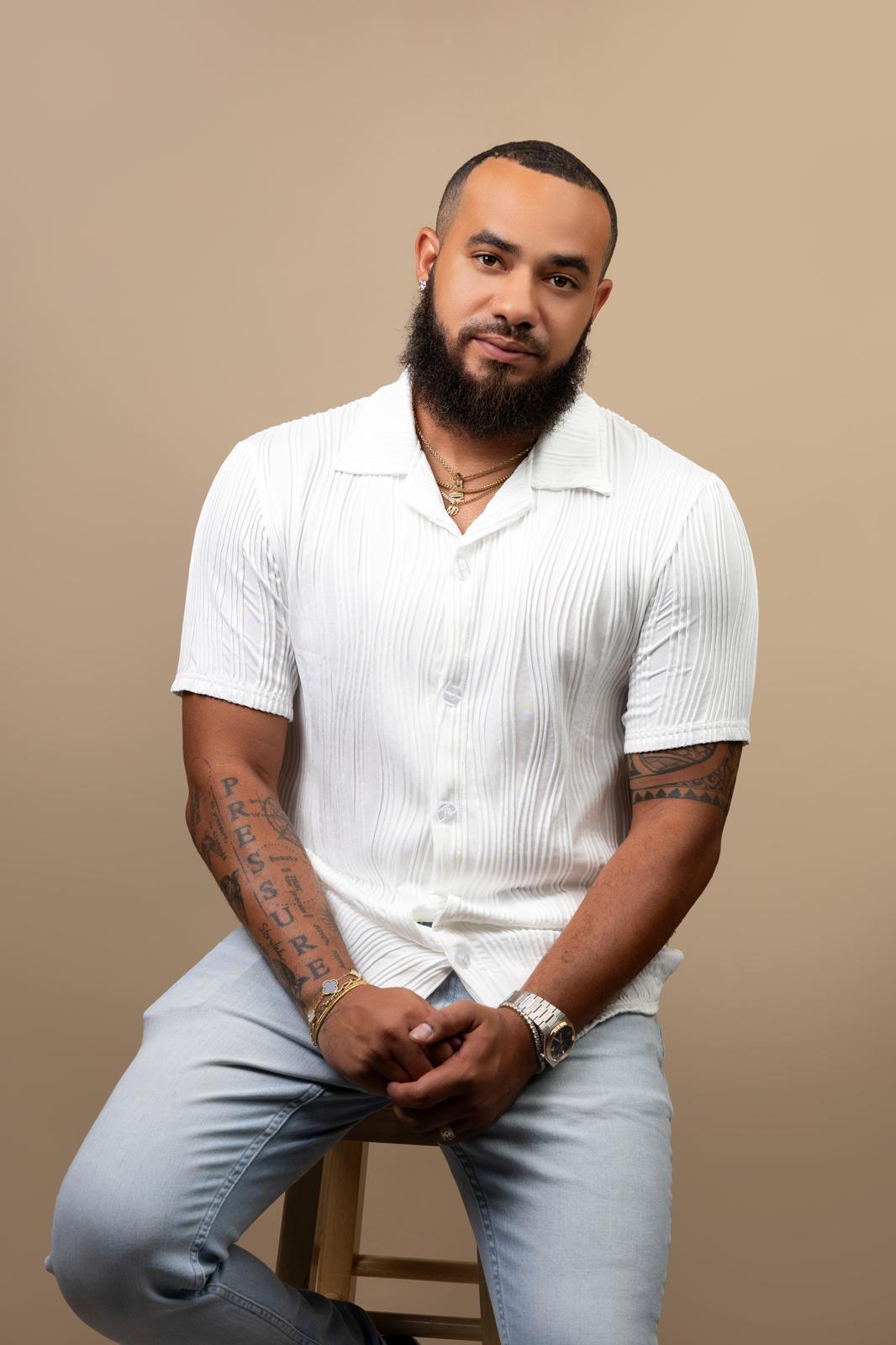 Man with a beard and tattoos, wearing a white shirt, sitting on a stool against a beige background.