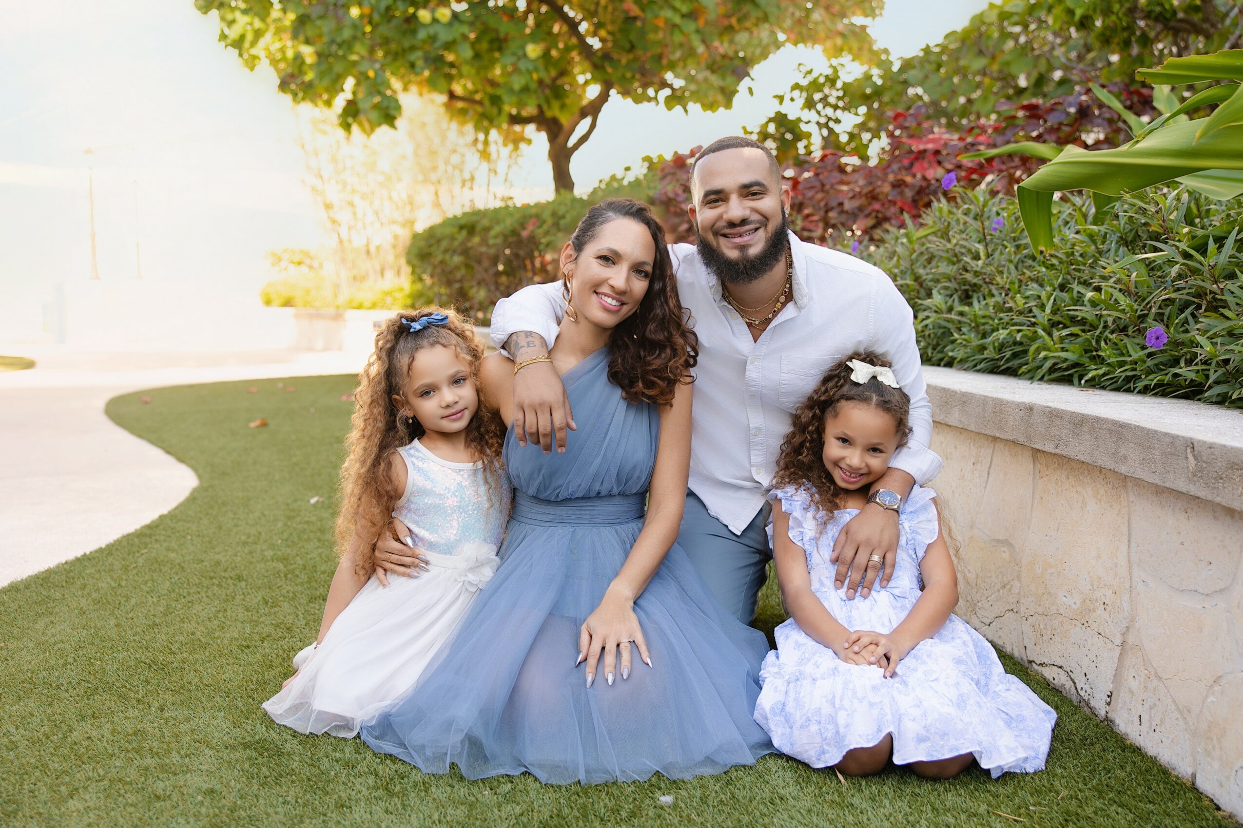 Family of four sitting outdoors on grass, smiling, with trees and plants in background.