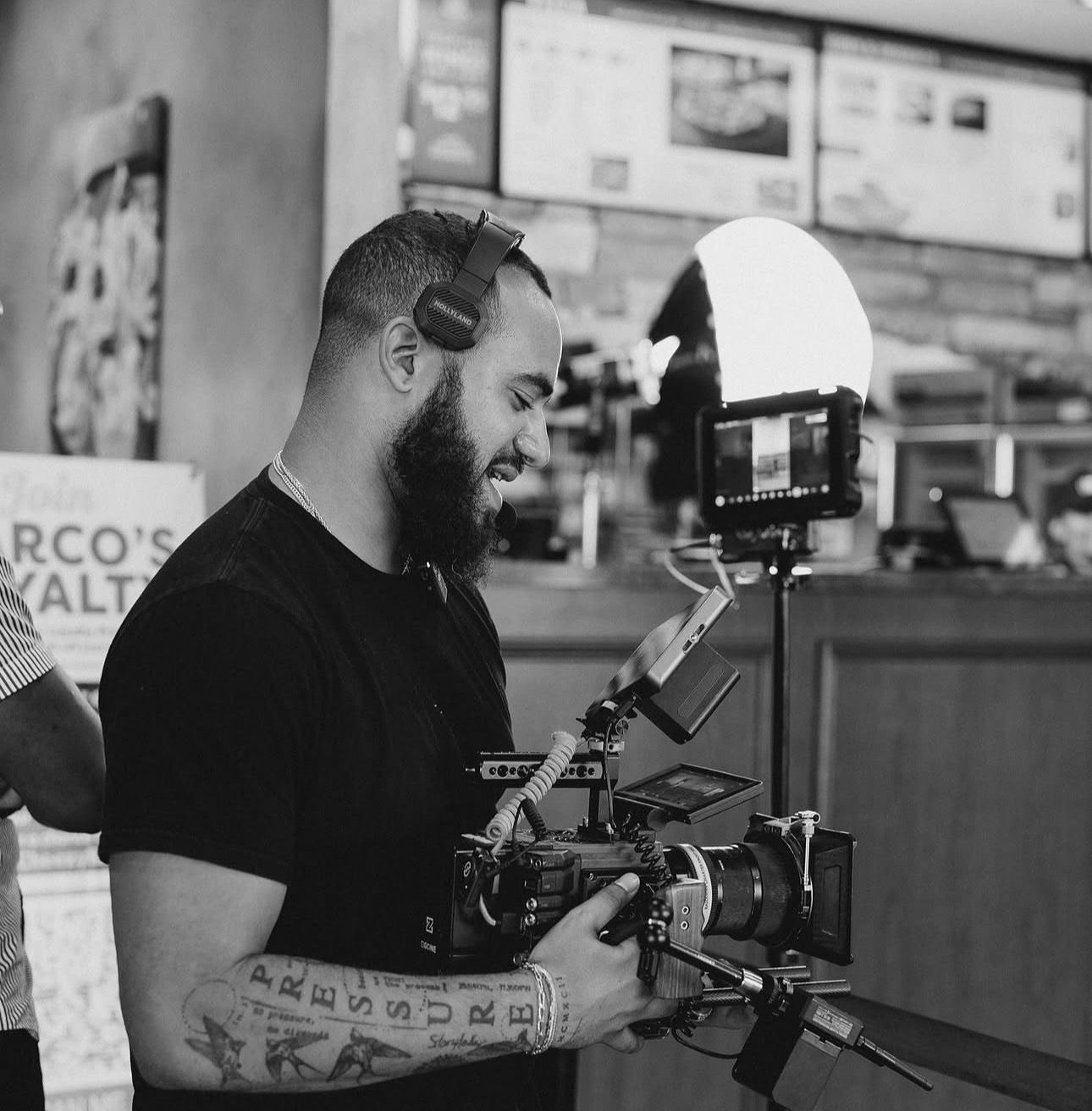 Man with a beard and tattoos wearing headphones, operating a camera in a fast-food restaurant, smiling, with menu screens in background.