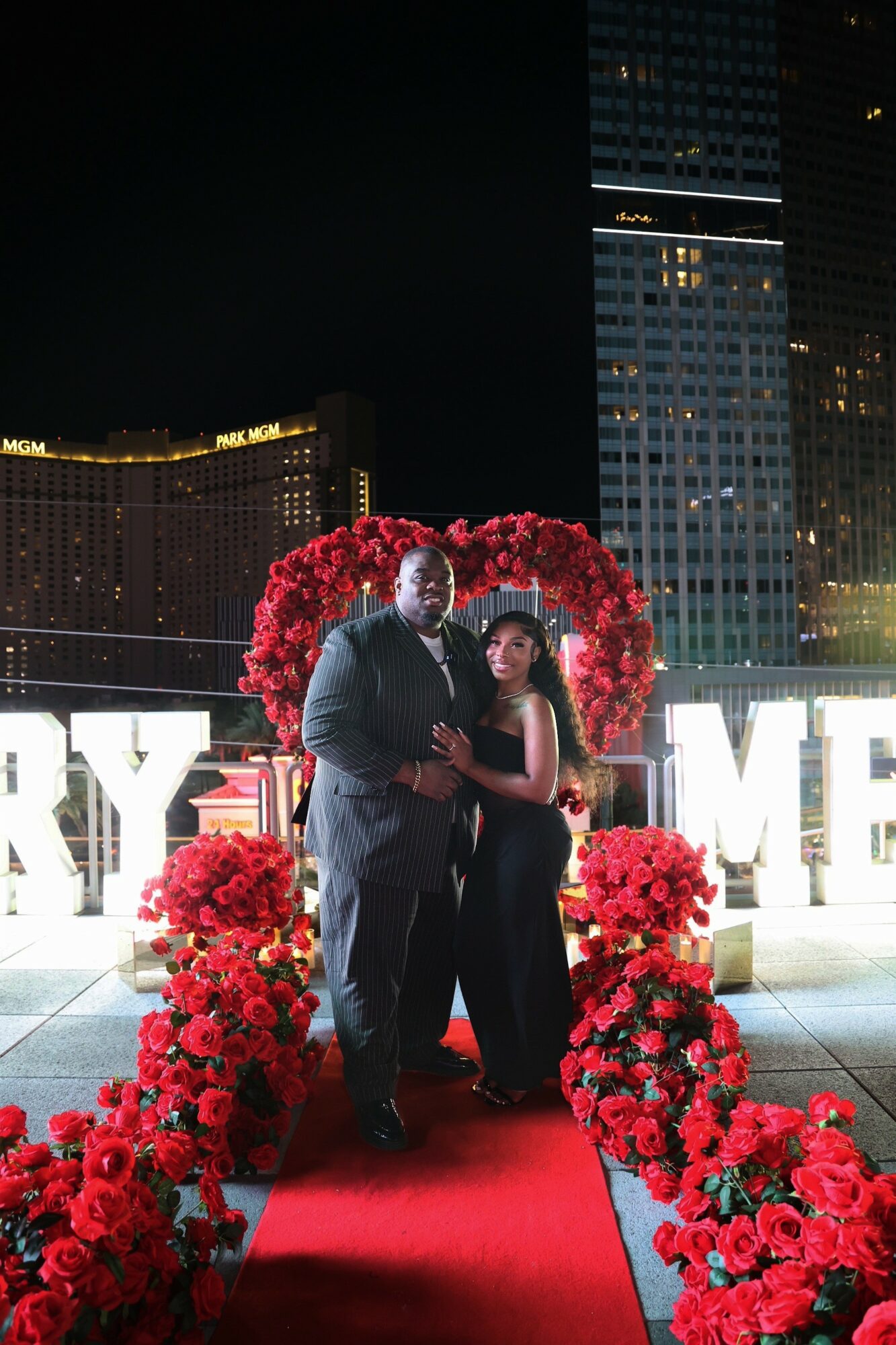 A couple stands on a red carpet with a heart-shaped floral arch behind them, city buildings in the background.