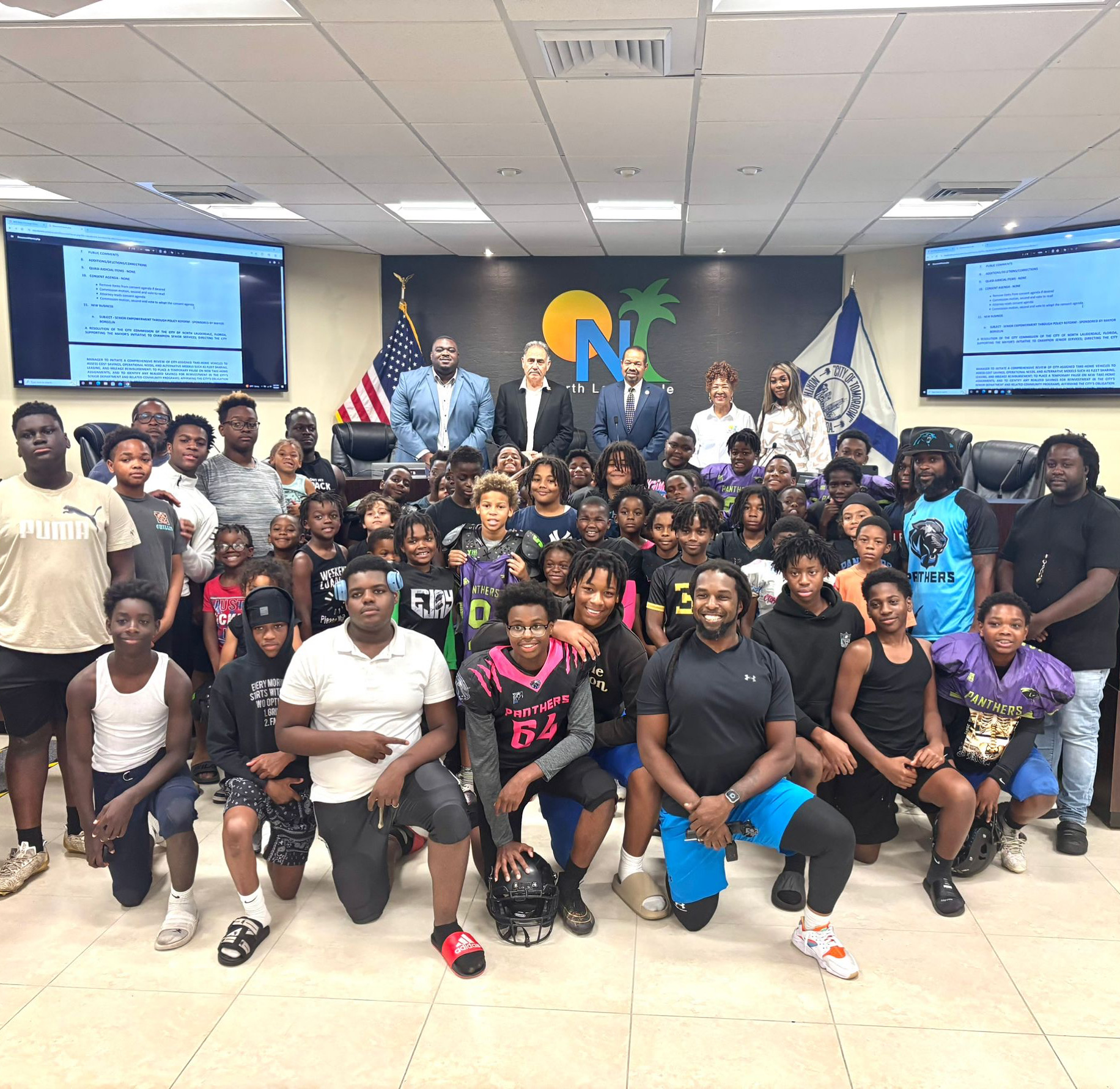 Group of people, including children and adults, gathered in a room with screens and flags, posing for a photo.