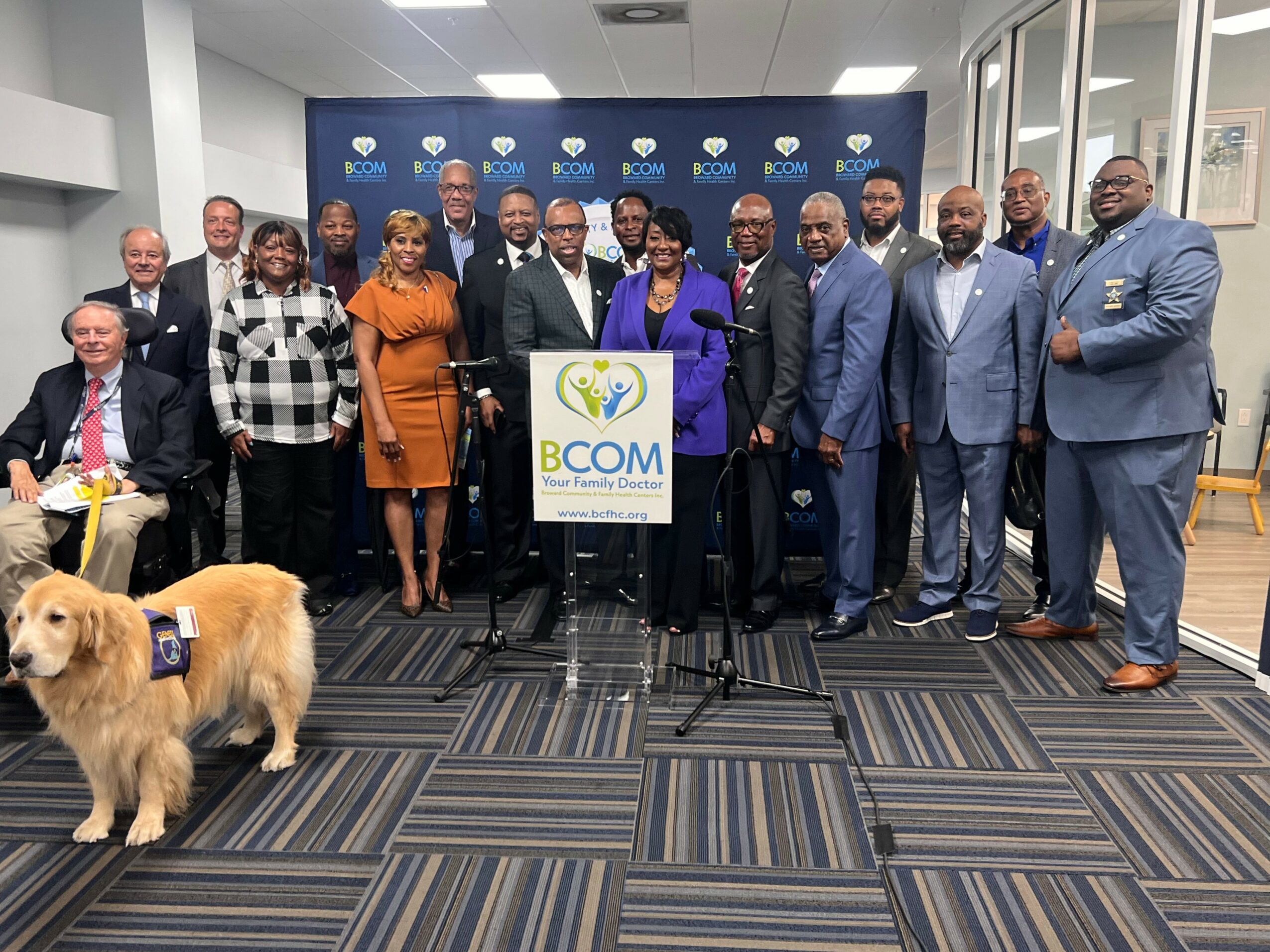 Group of people standing in a room with a dog in the foreground, a banner, and a sign that reads 'BCOM'.