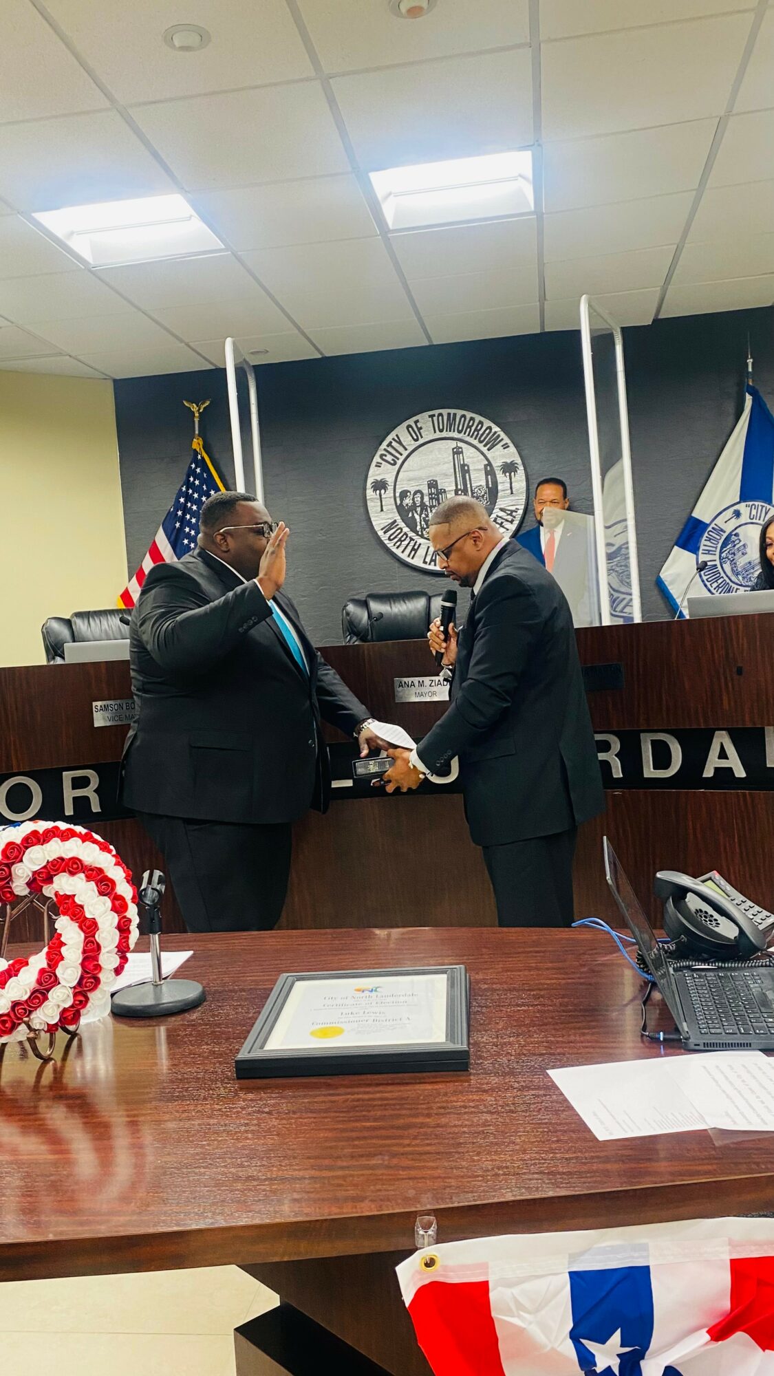 Two men in suits shake hands in a formal setting with flags and a seal behind them, a table with documents in foreground.