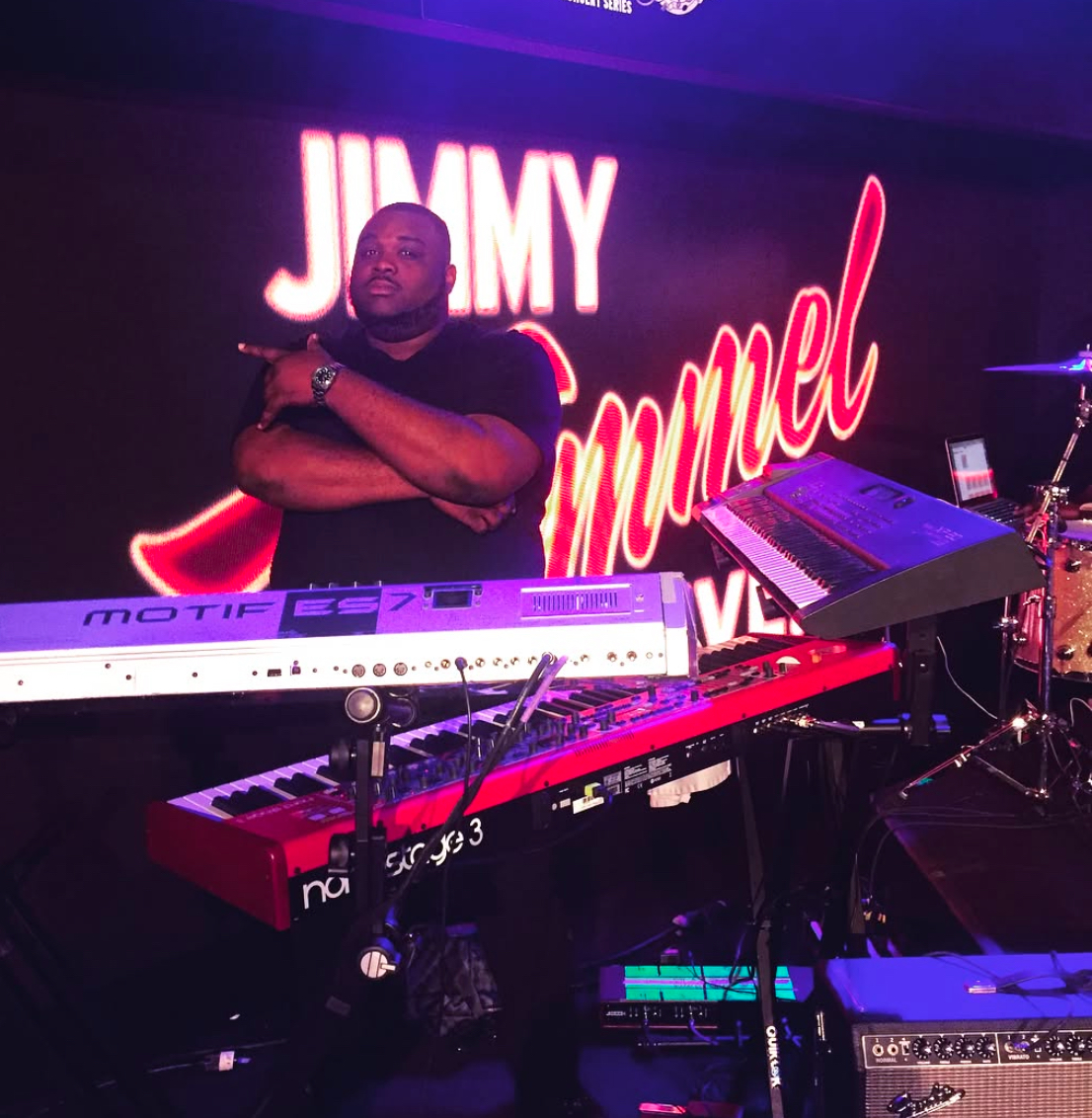 A man stands behind a keyboard setup on stage with a neon sign in the background reading 'JIMMY' and 'Mummel'.