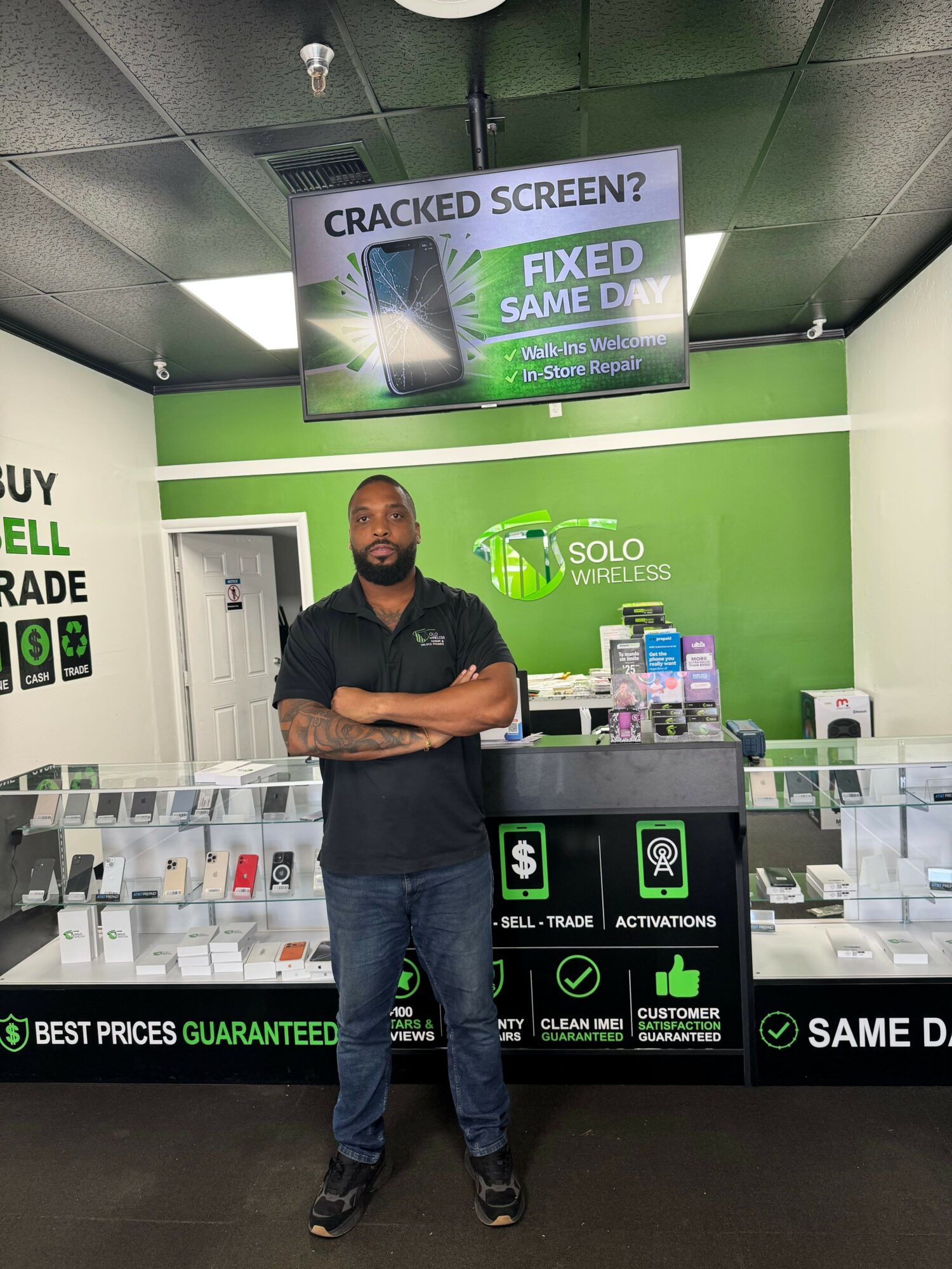 Man standing inside a store with green and black walls, digital screen, and display cases, arms crossed, smiling.