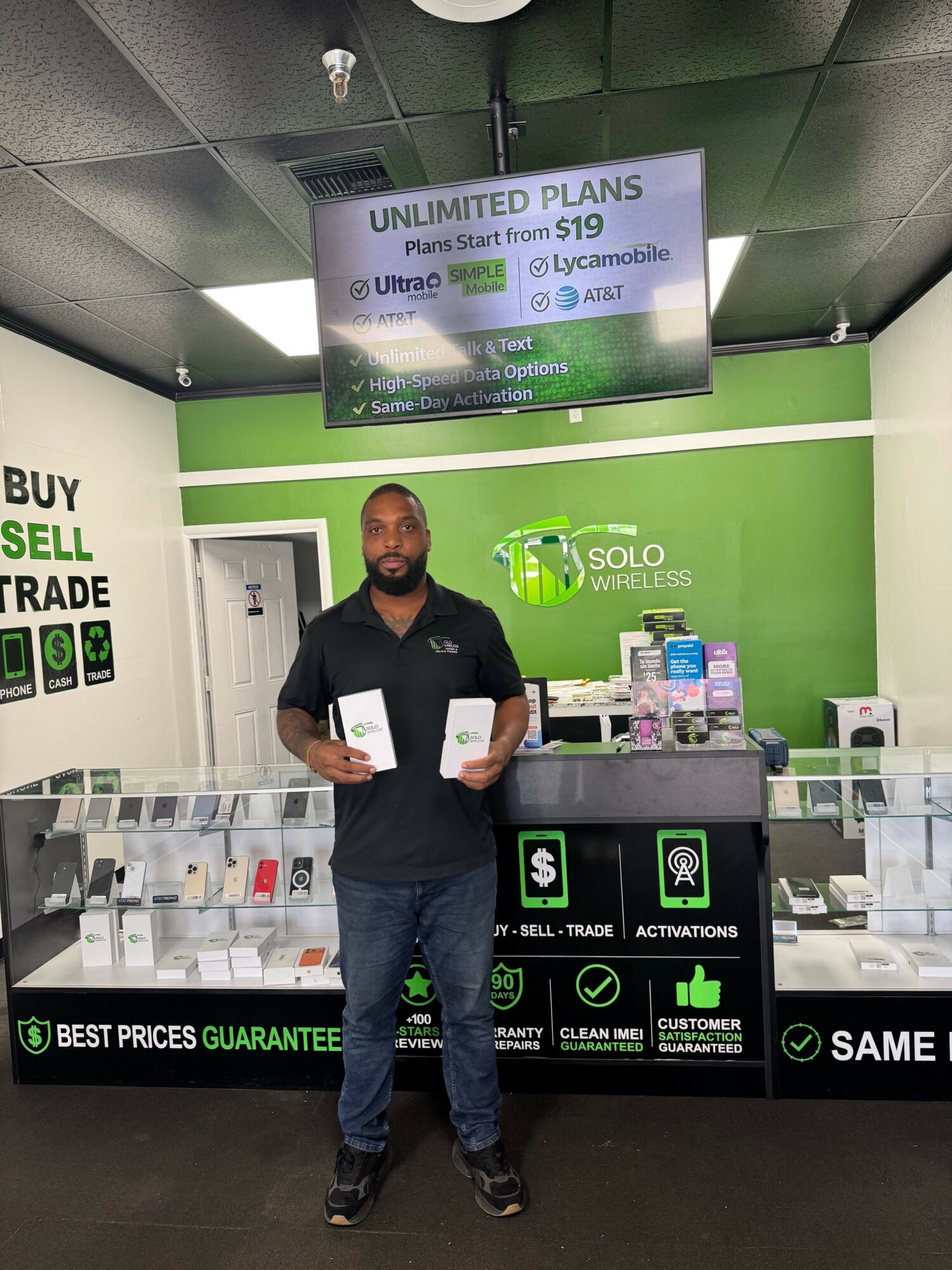 Man standing inside a store holding a laptop, with a green and black display behind him, and a digital screen overhead.
