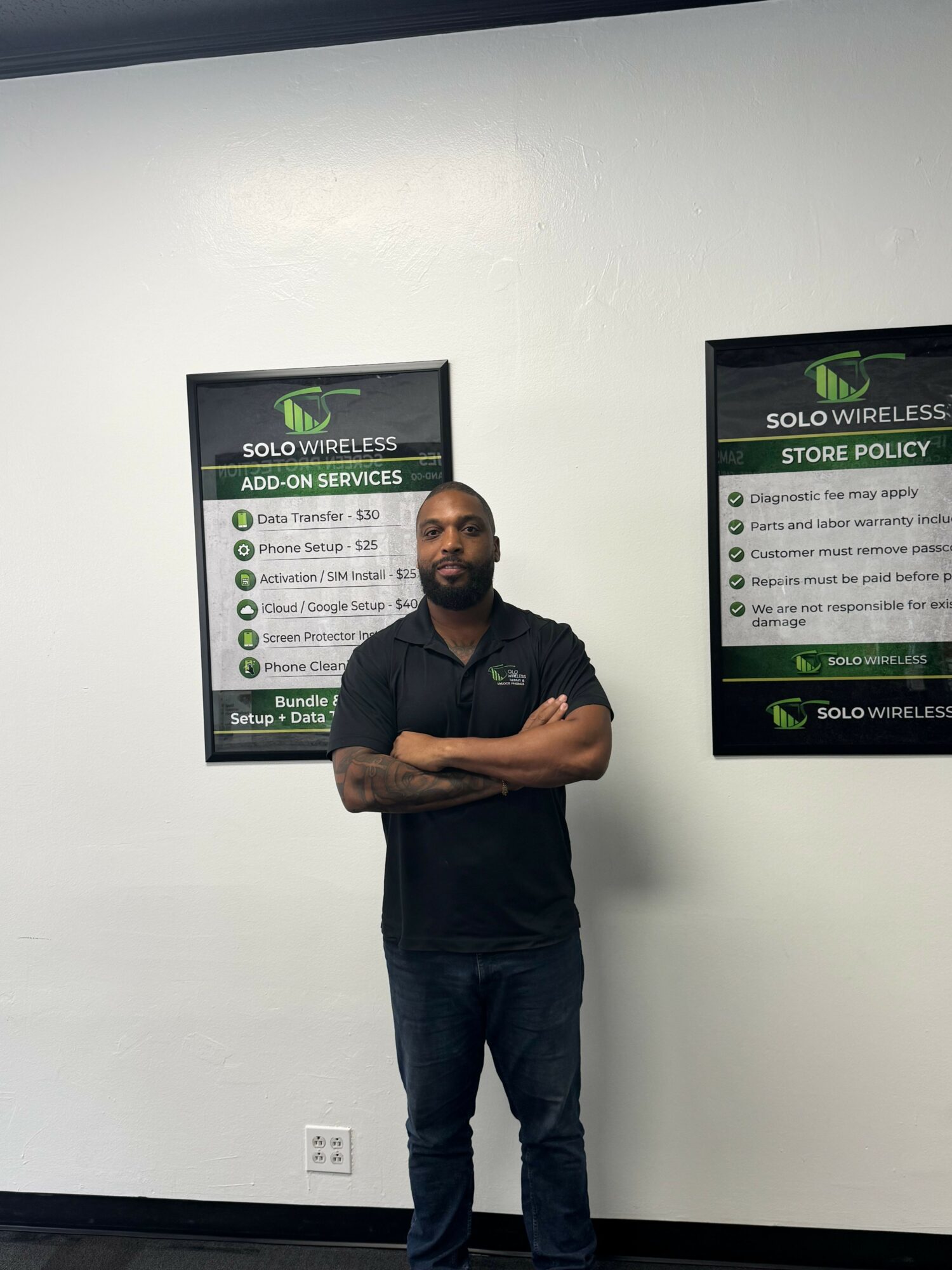 Man standing with arms crossed in front of a white wall with informational posters, facing camera.