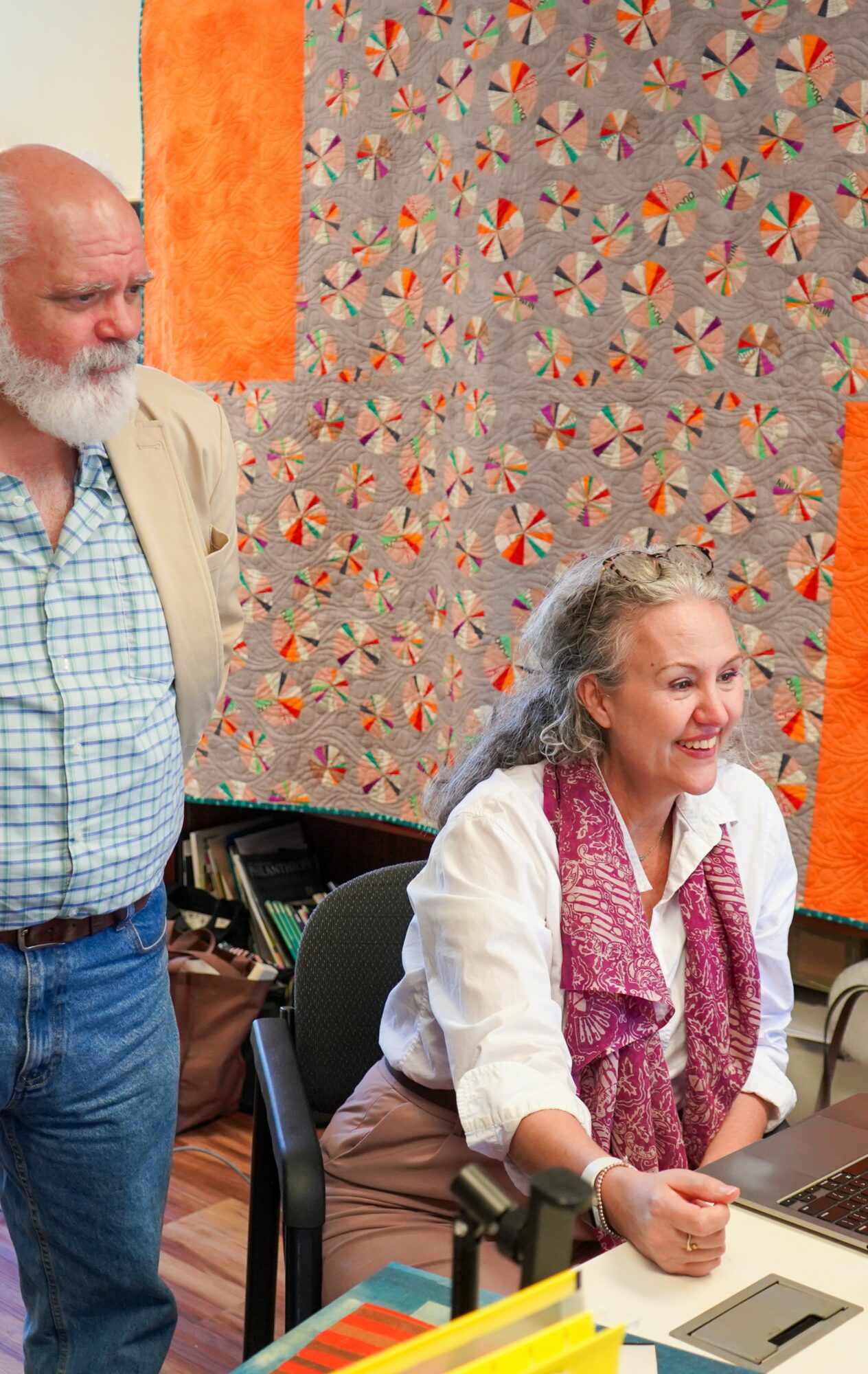 An elderly man and woman are in a room with colorful, patterned wall art. The woman is sitting at a table with a laptop.