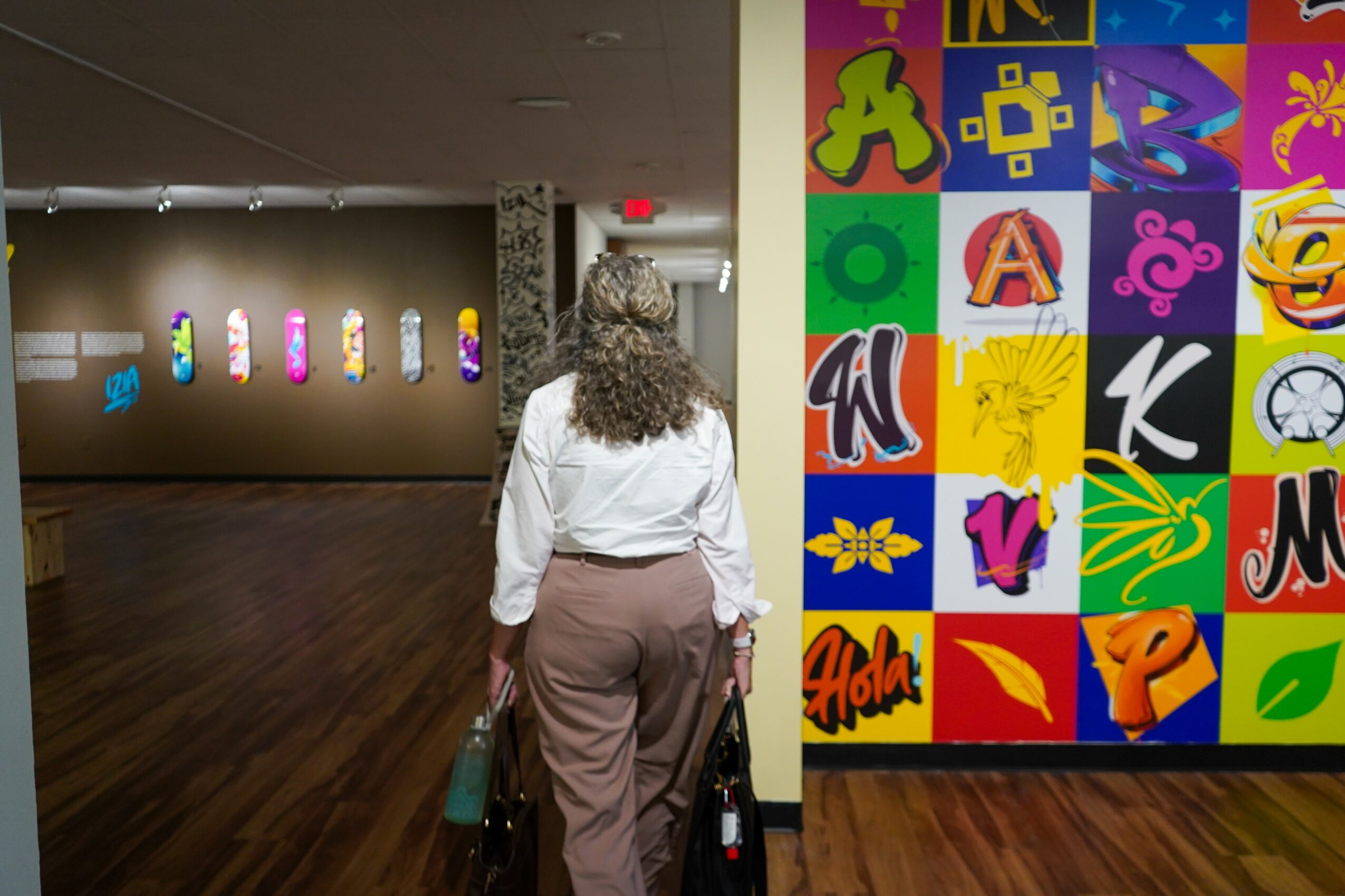Person with curly hair wearing a white shirt and beige pants stands in an art gallery, looking at colorful artwork on the wall.