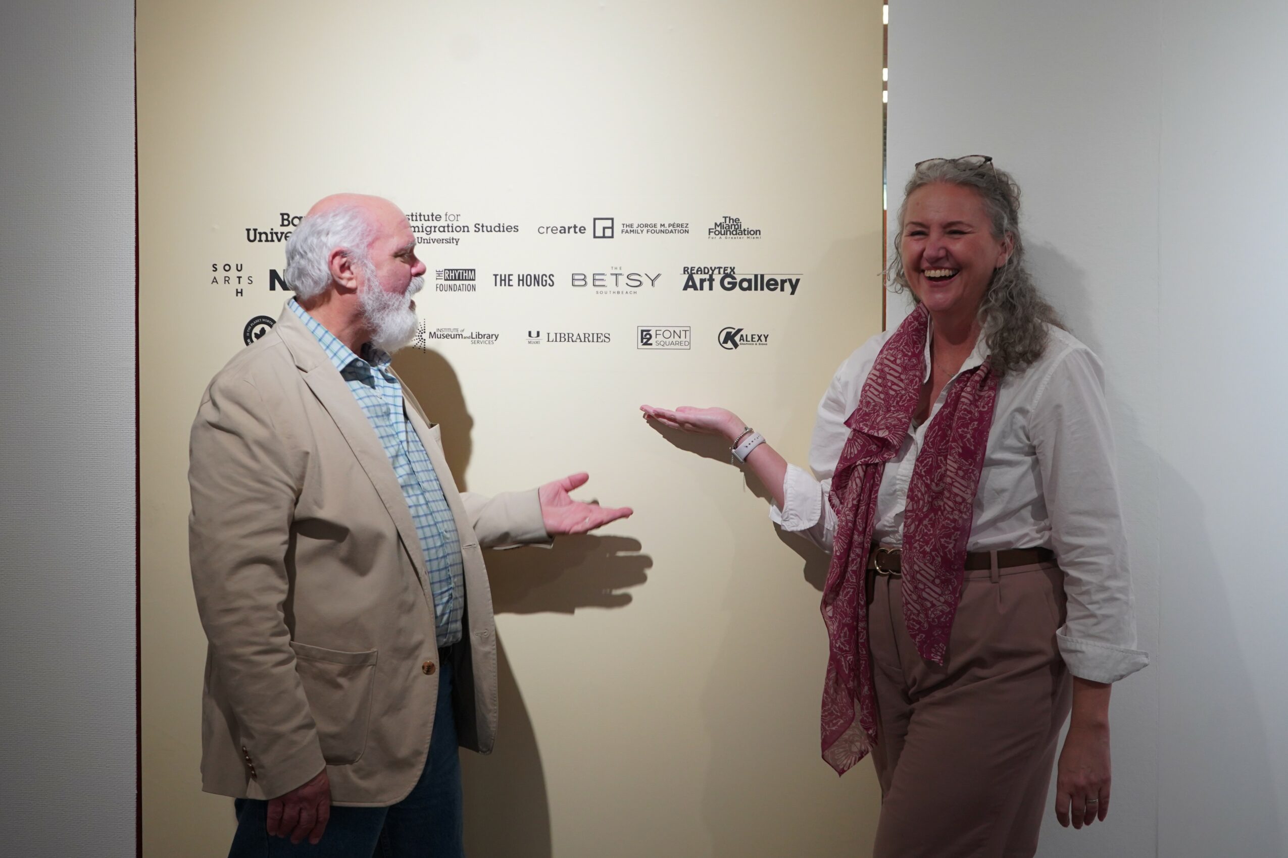 Two people, a man and a woman, are smiling and talking in front of a beige wall with logos. The woman is holding a small object.