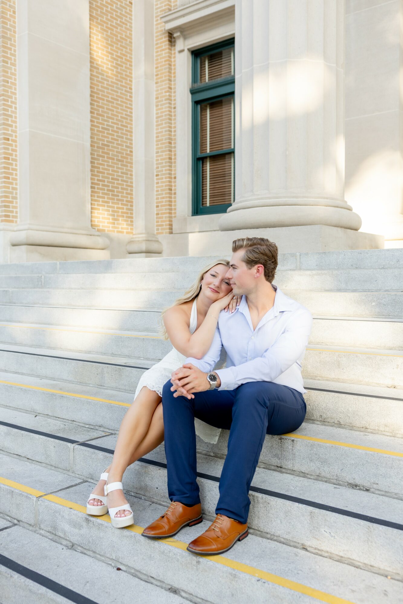 A couple sitting on steps outside a building, smiling and leaning close to each other, with columns and a window behind them.