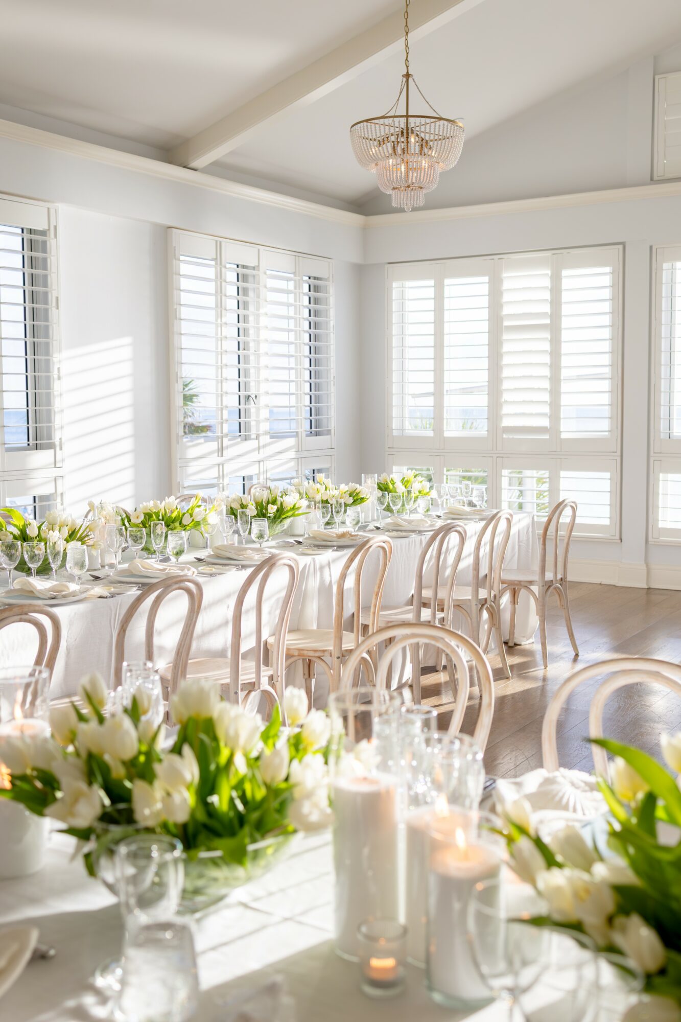 Elegant dining room with large windows, white chairs, floral centerpieces, and candles on tables, bright and airy atmosphere.