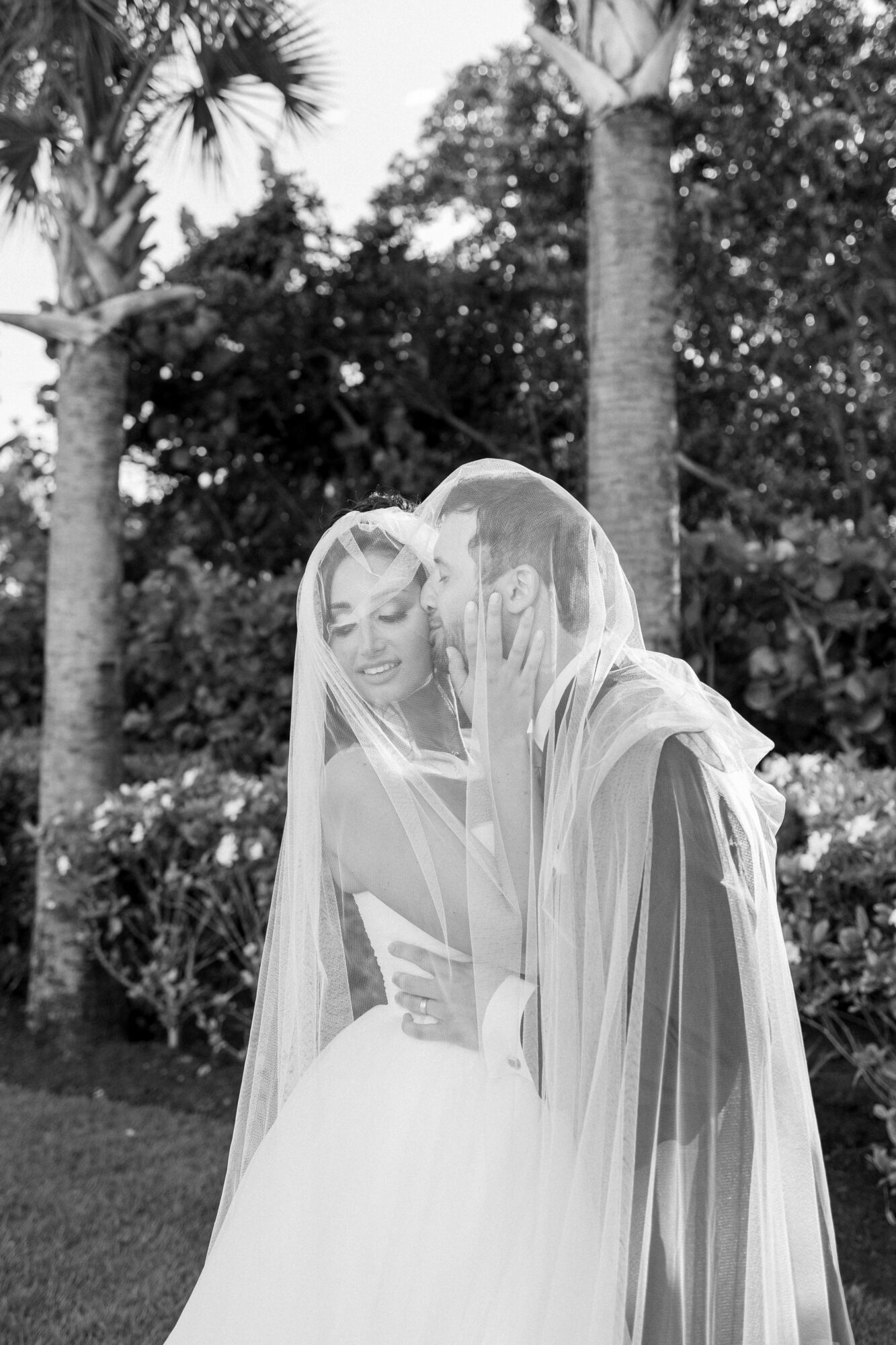 Two women in wedding dresses with veils, smiling and close together outdoors, trees in background.