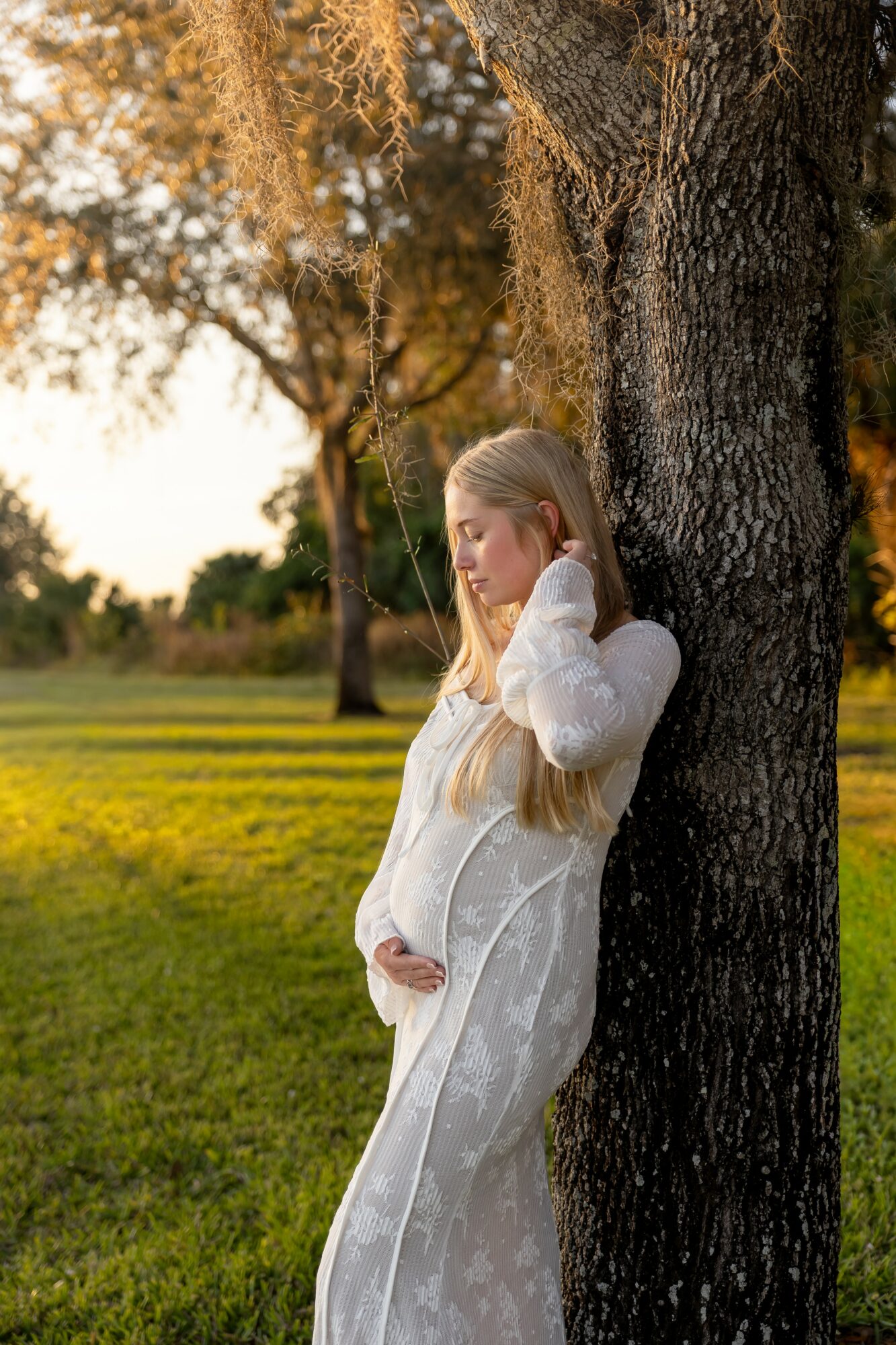 Pregnant woman with long blonde hair leaning against a tree outdoors during sunset, holding her belly gently.