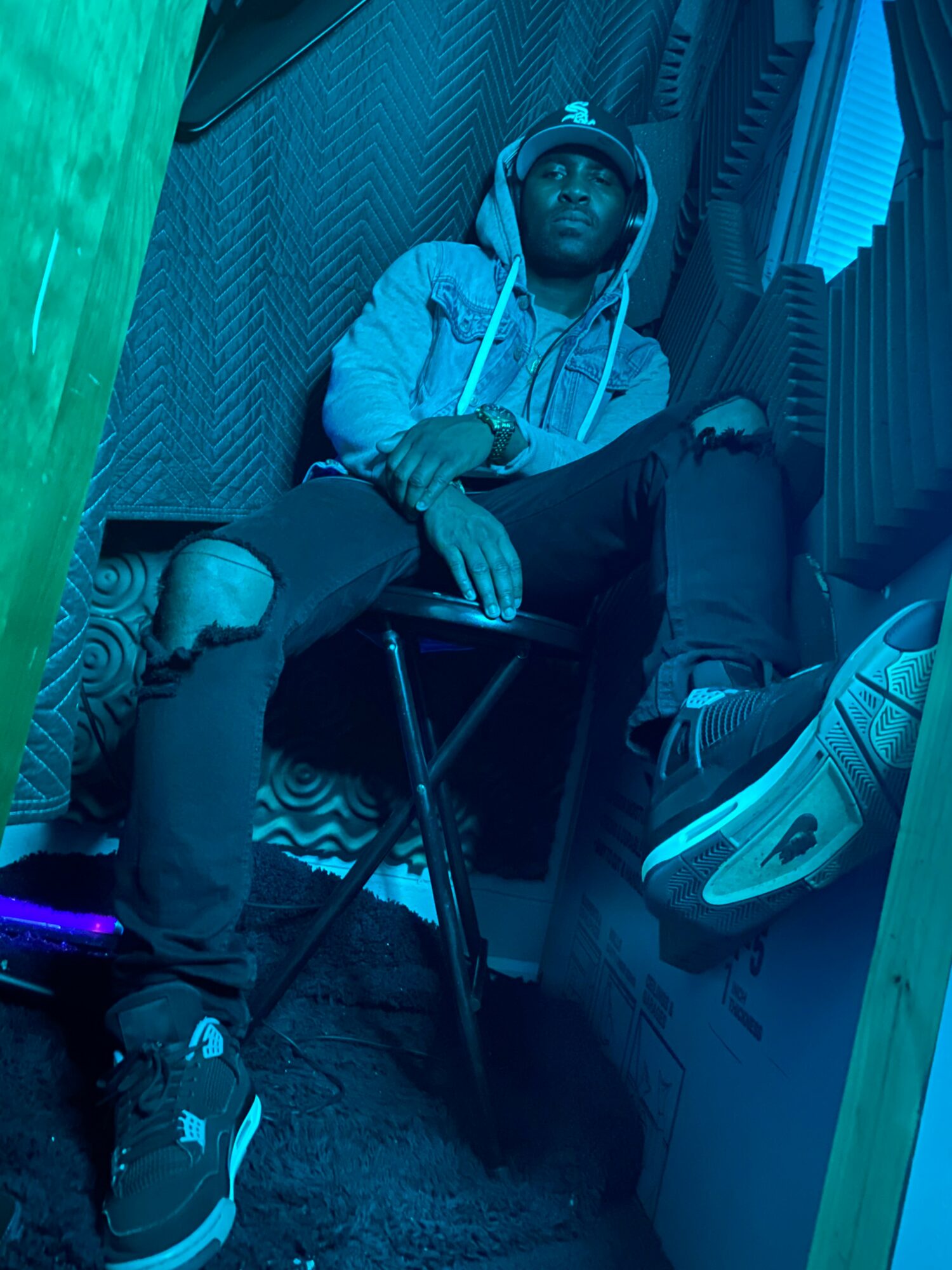 Young man sitting on a chair, wearing a hoodie, ripped jeans, and sneakers, in a room with blue lighting.