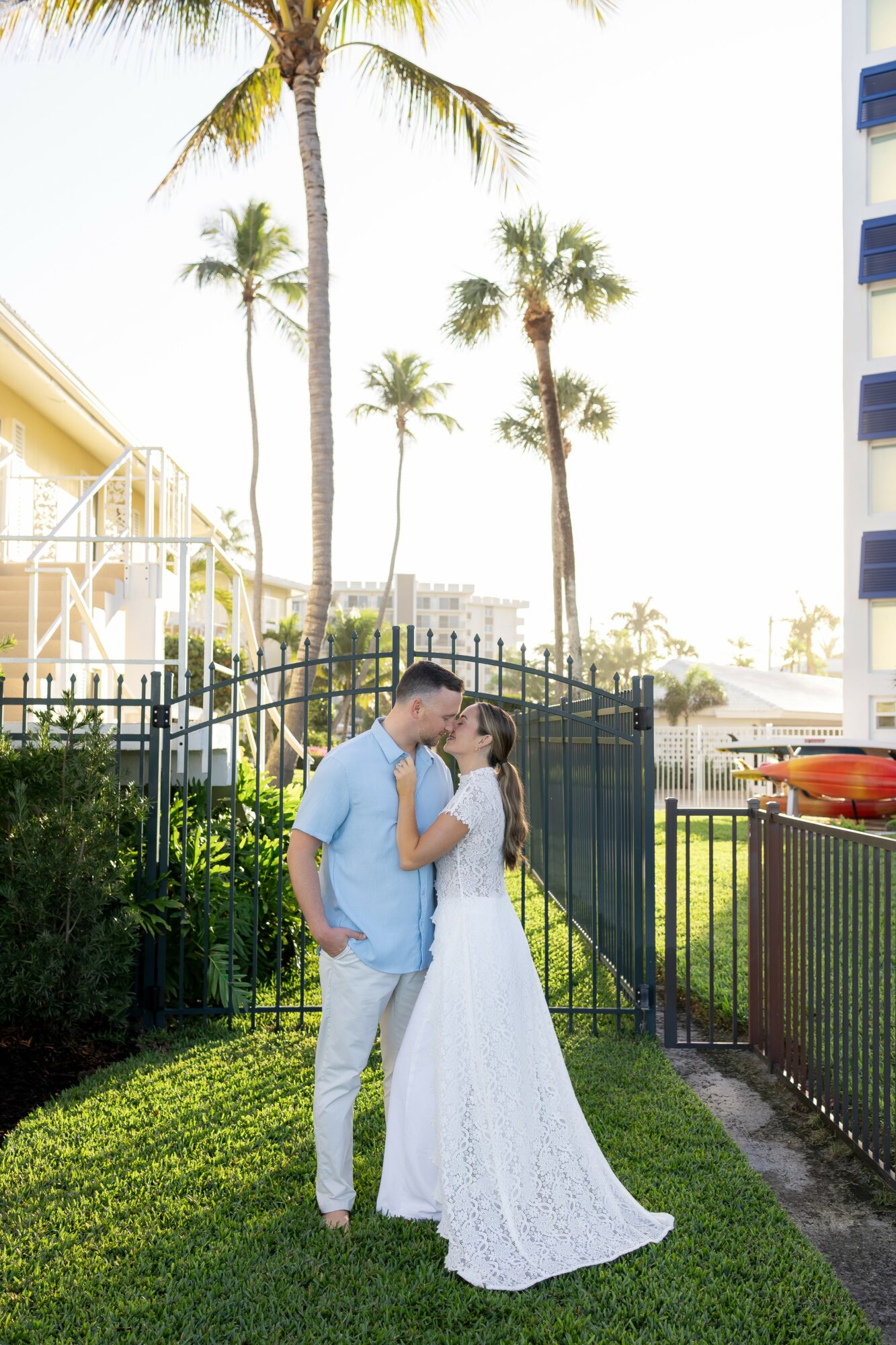 A couple in wedding attire kissing outdoors with palm trees and buildings in the background.