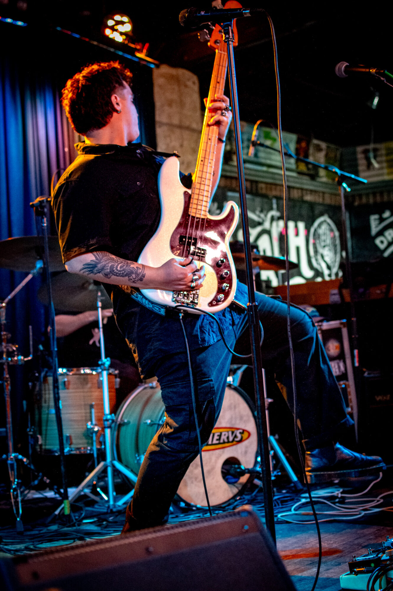 Musician playing electric guitar on stage with drums and microphone, stage lights in background.