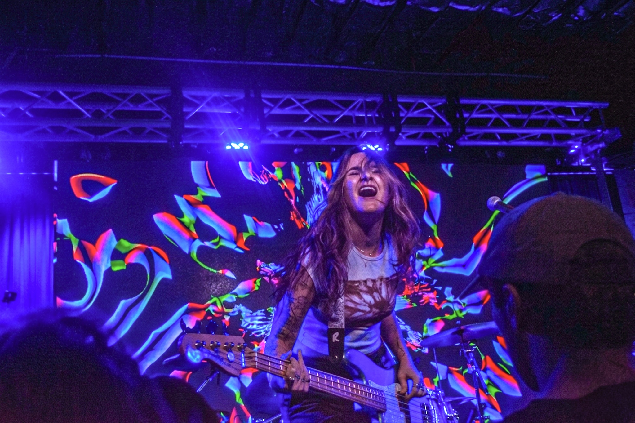 A woman with long hair plays an electric guitar on stage with colorful lights and abstract visuals behind her.