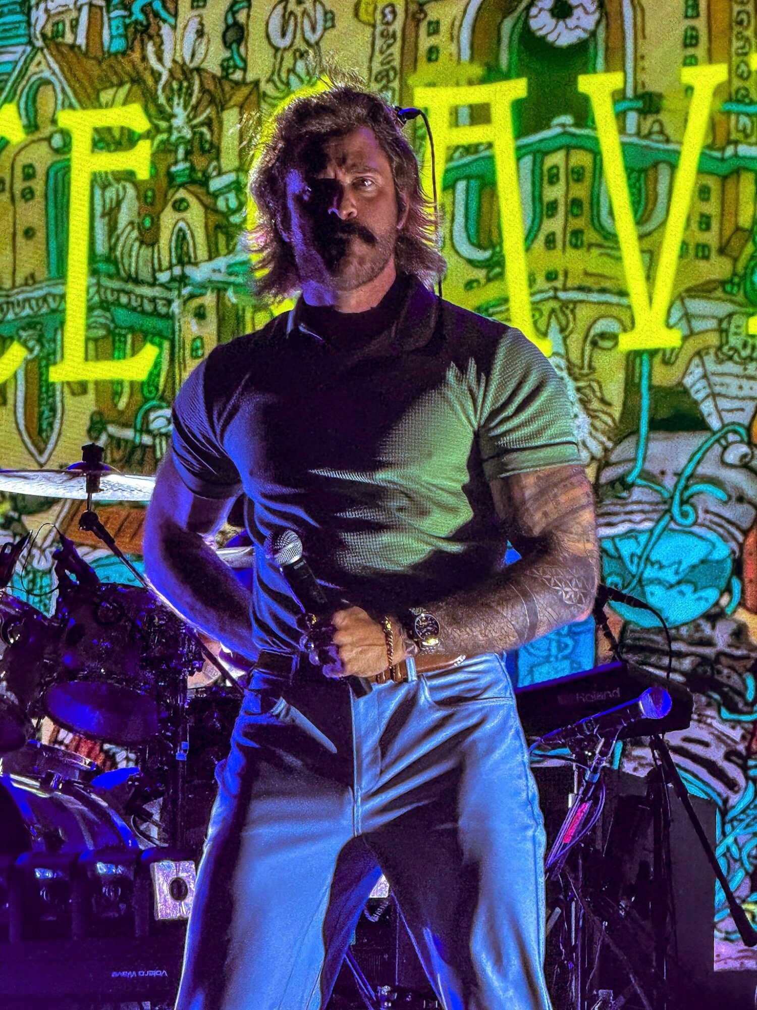 Man with long hair and beard stands in front of colorful graffiti background, wearing a black and green shirt and blue pants.
