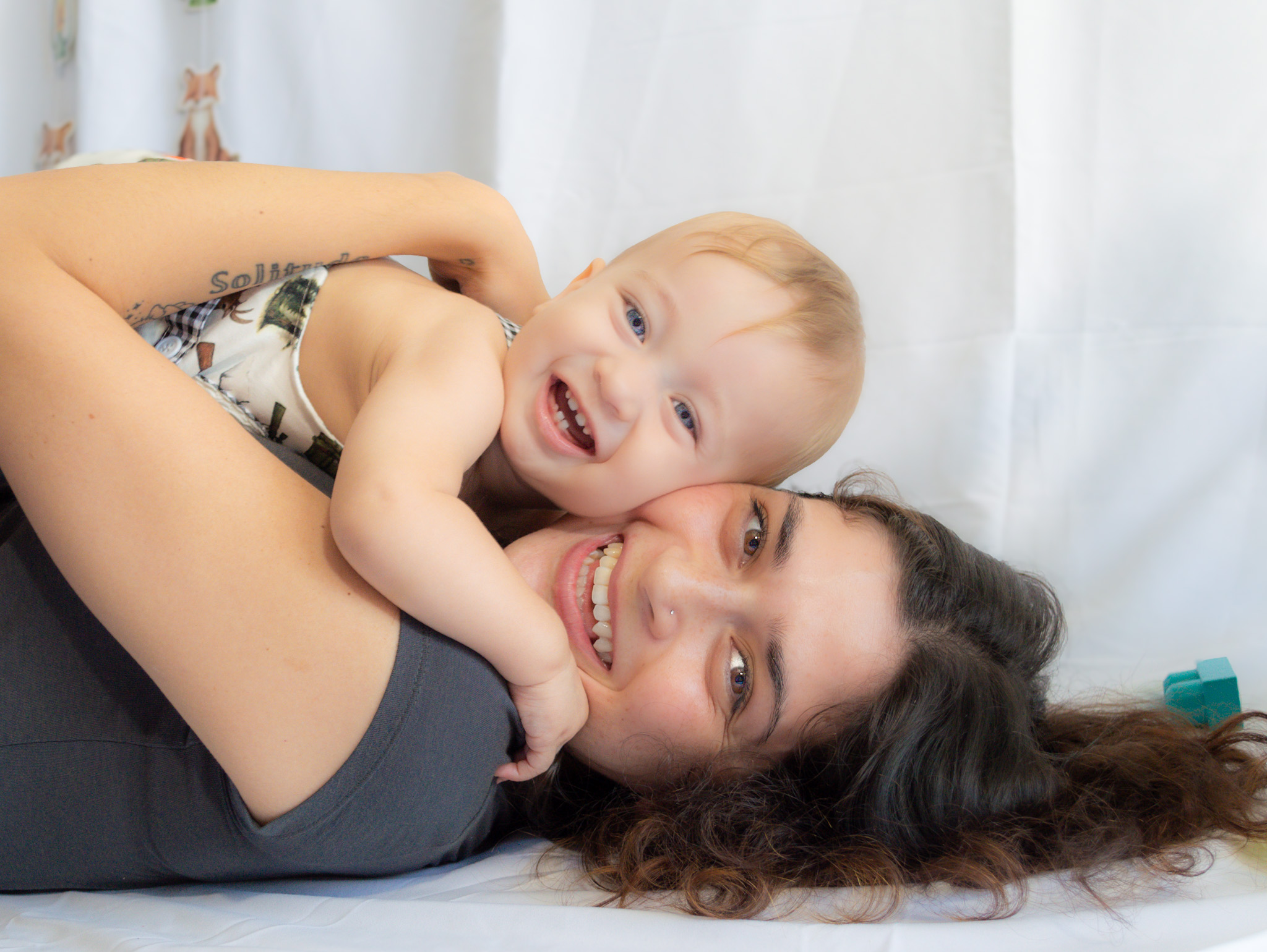 A woman lying on her back with a young child on her chest, both smiling, in a bright room with white curtains.