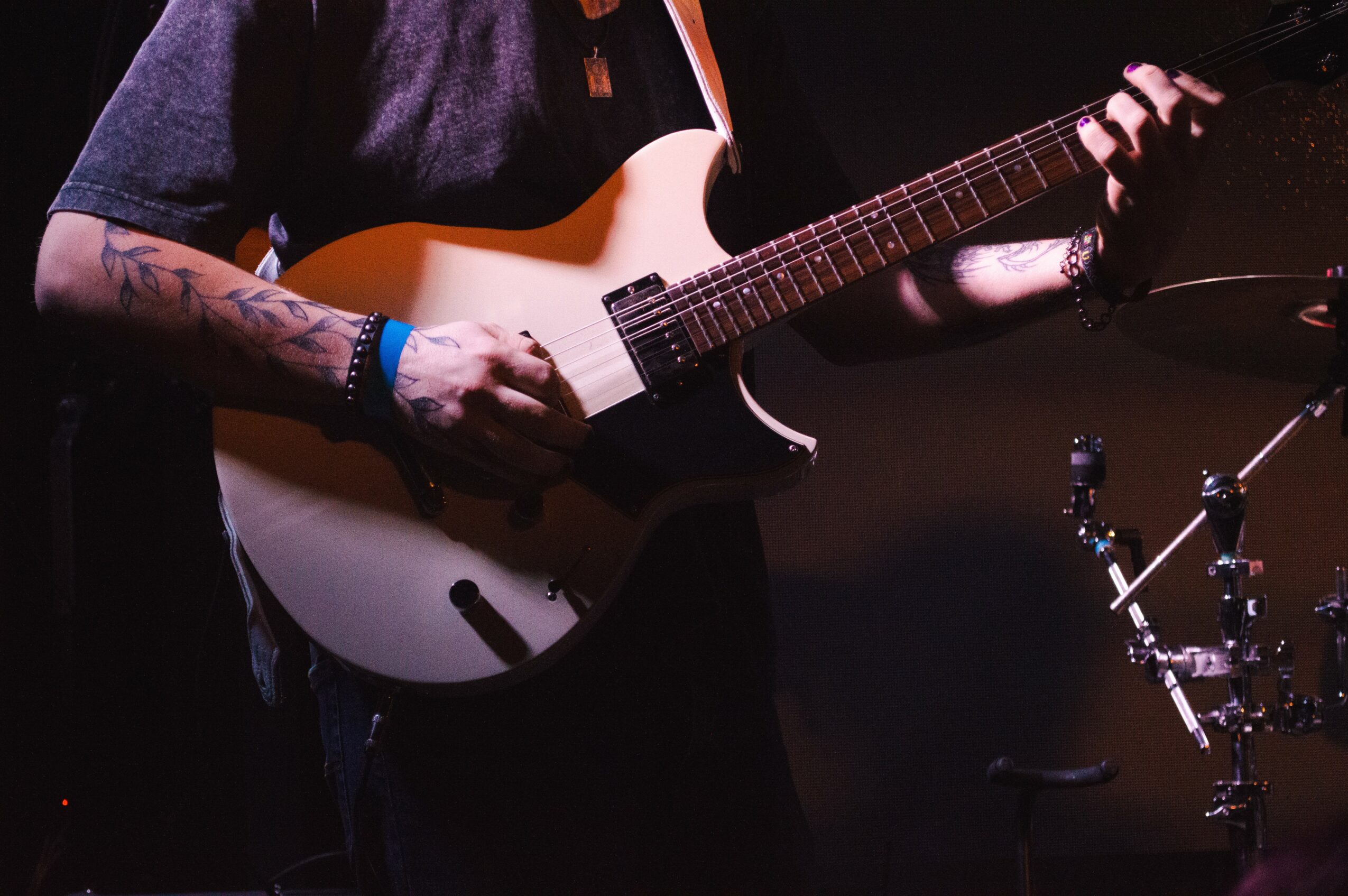 Person playing an electric guitar with tattoos on arms, dark background, stage lighting, and drum set nearby.