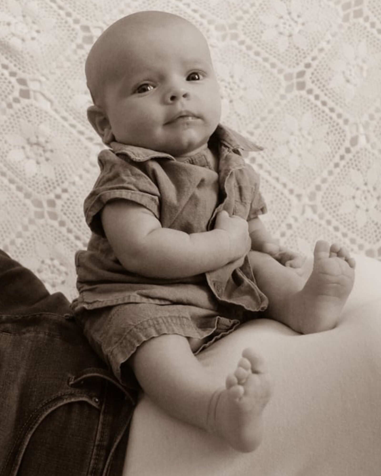 Baby sitting on a person's lap, looking at the camera, with a lace background, wearing a short-sleeved outfit.