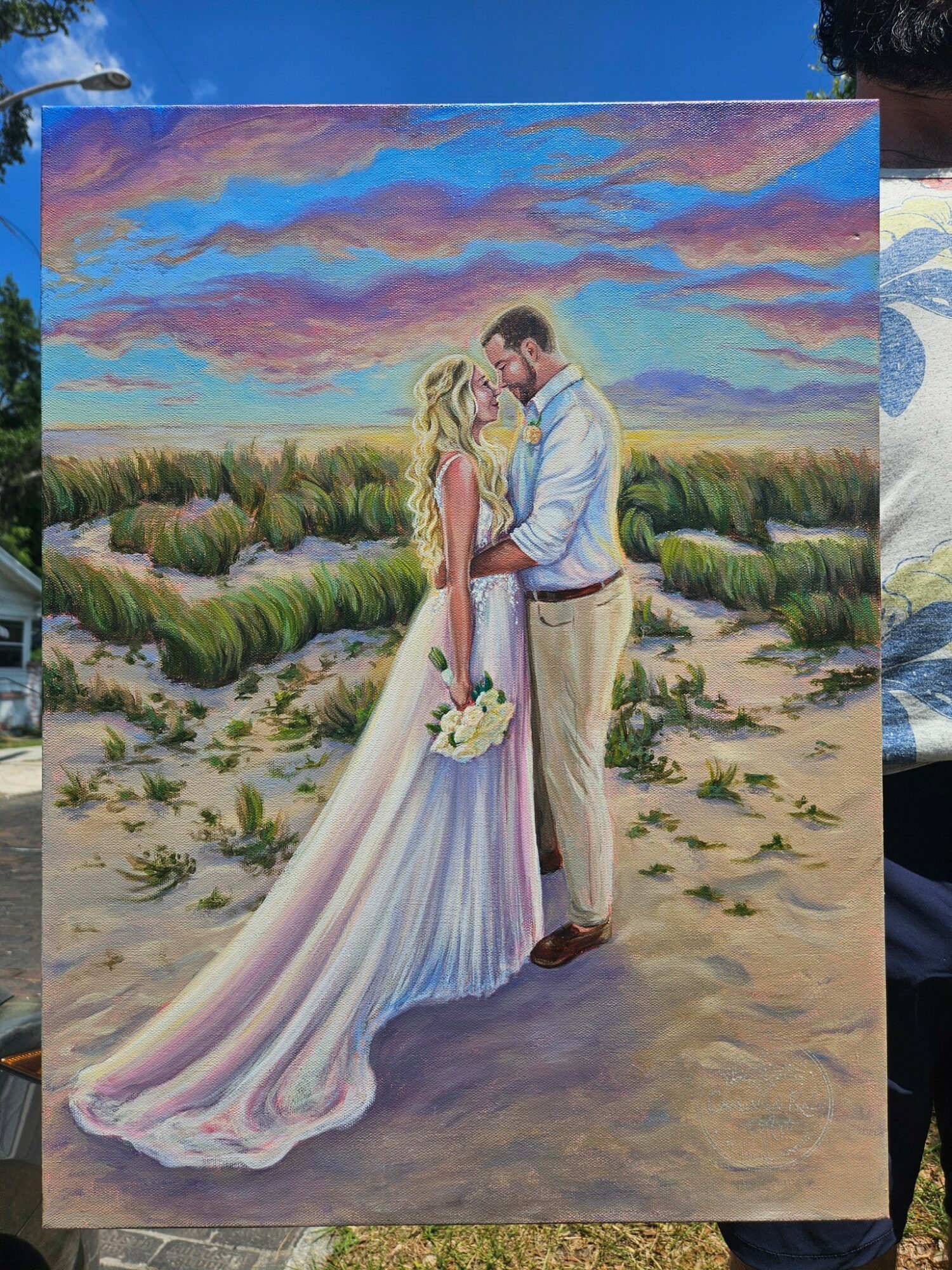 A bride and groom stand close together outdoors, holding hands, with a colorful sky and landscape in the background.