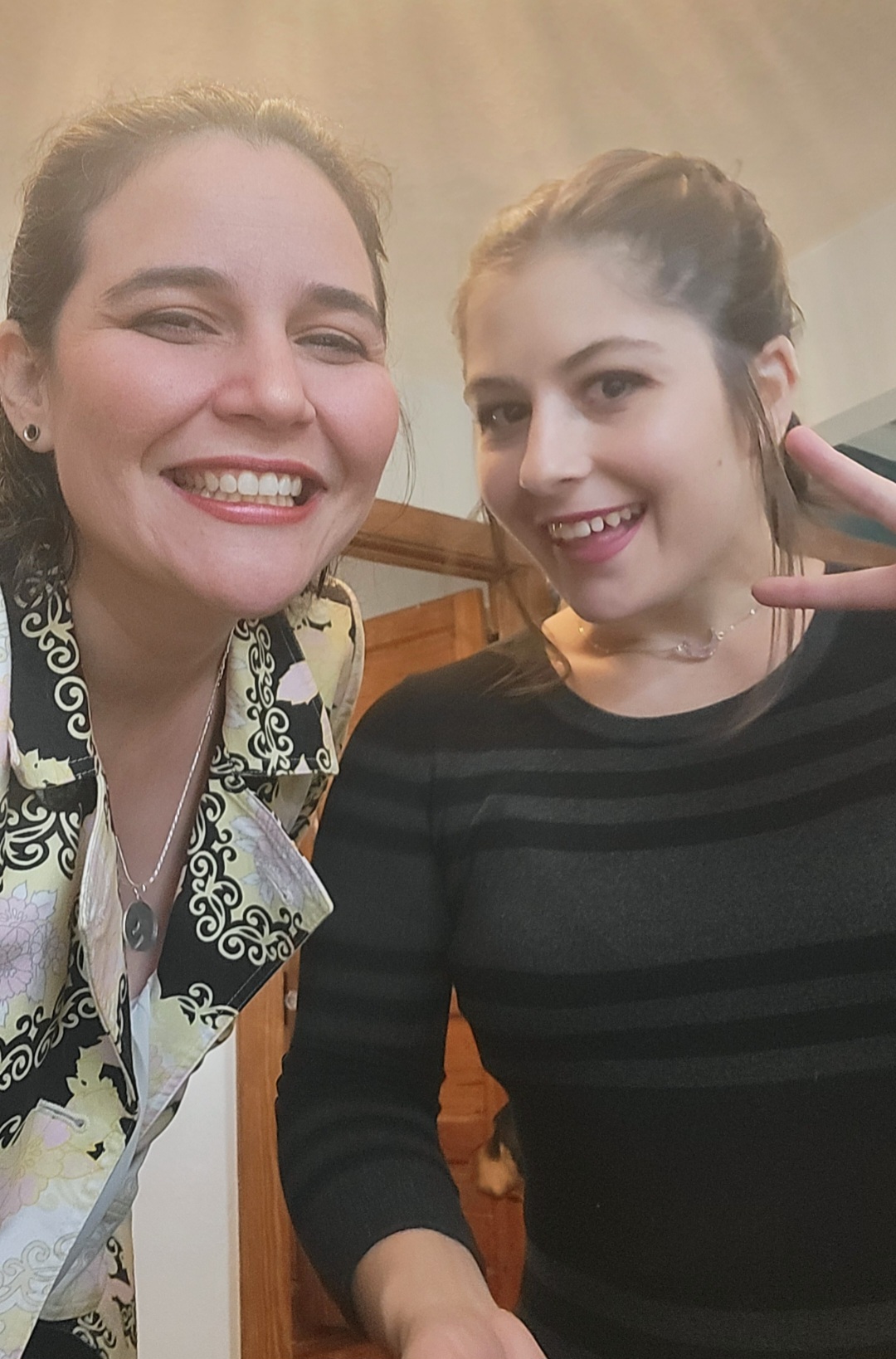 Two smiling women taking a selfie indoors, one making a peace sign with her hand.