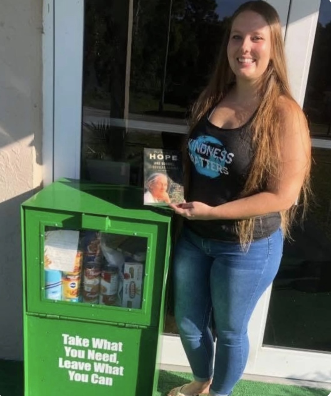 Young woman with long hair smiling, standing outside near a green donation box, holding a flyer, wearing a black tank top and jeans.