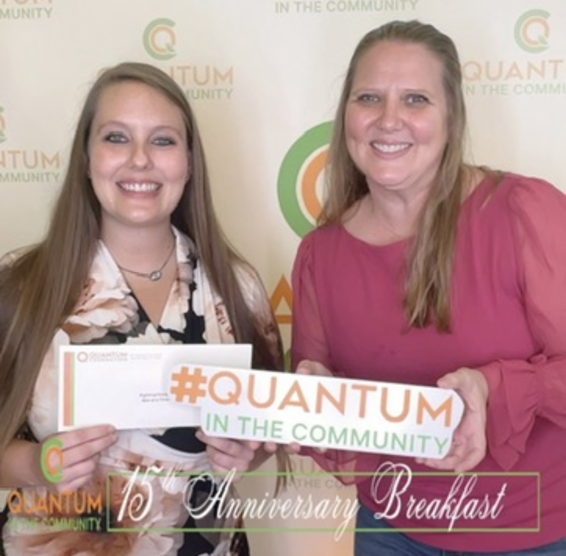 Two women smiling, holding a sign that says #QUANTUM IN THE COMMUNITY, standing in front of a backdrop with event logos.