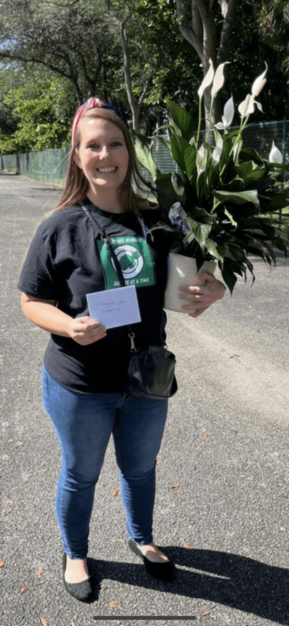 Woman holding a large plant with white flowers outdoors, smiling, wearing a black T-shirt and jeans.