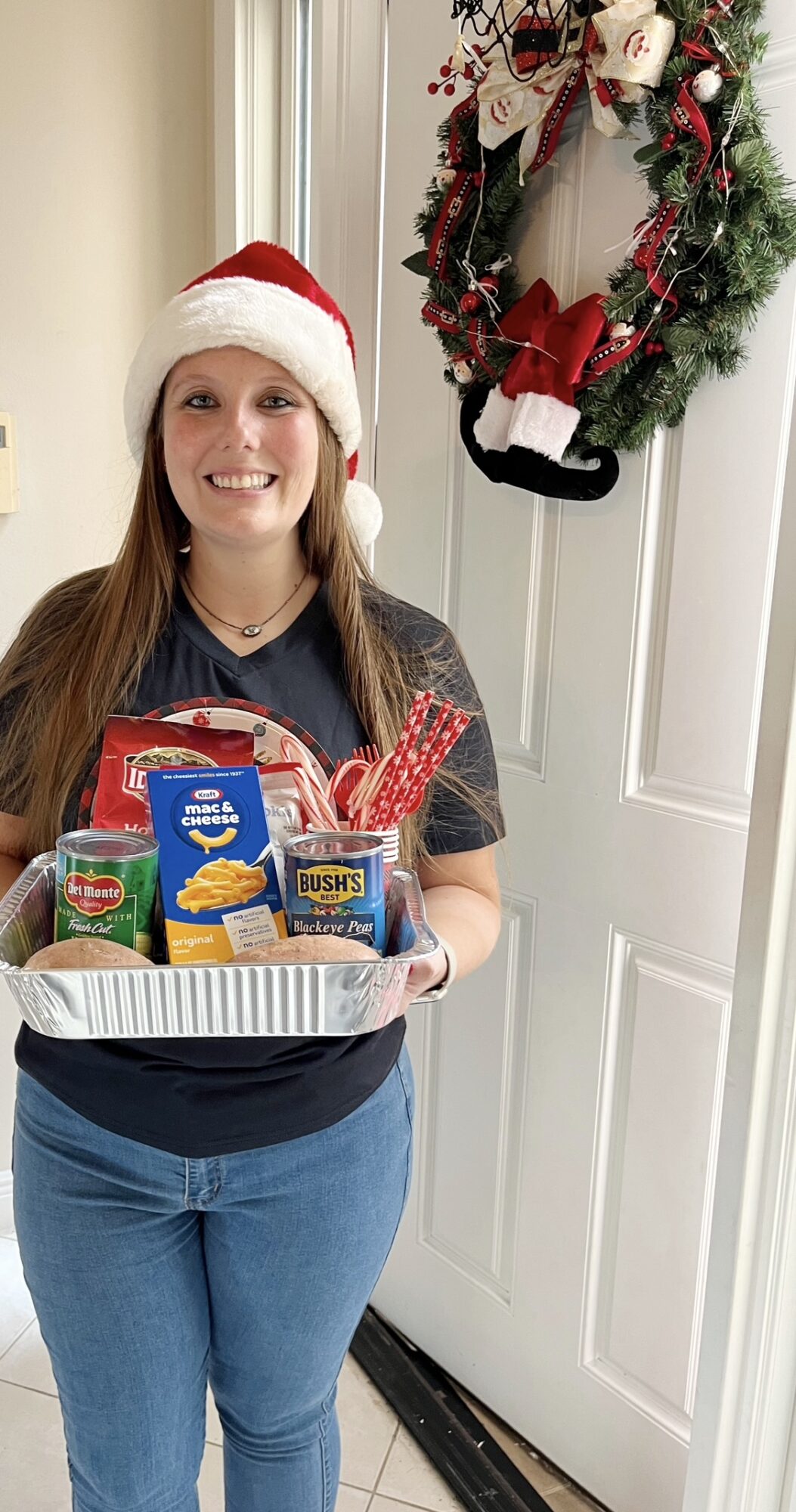 Young woman wearing a Santa hat holding a tray of holiday food, standing near a door with a Christmas wreath.