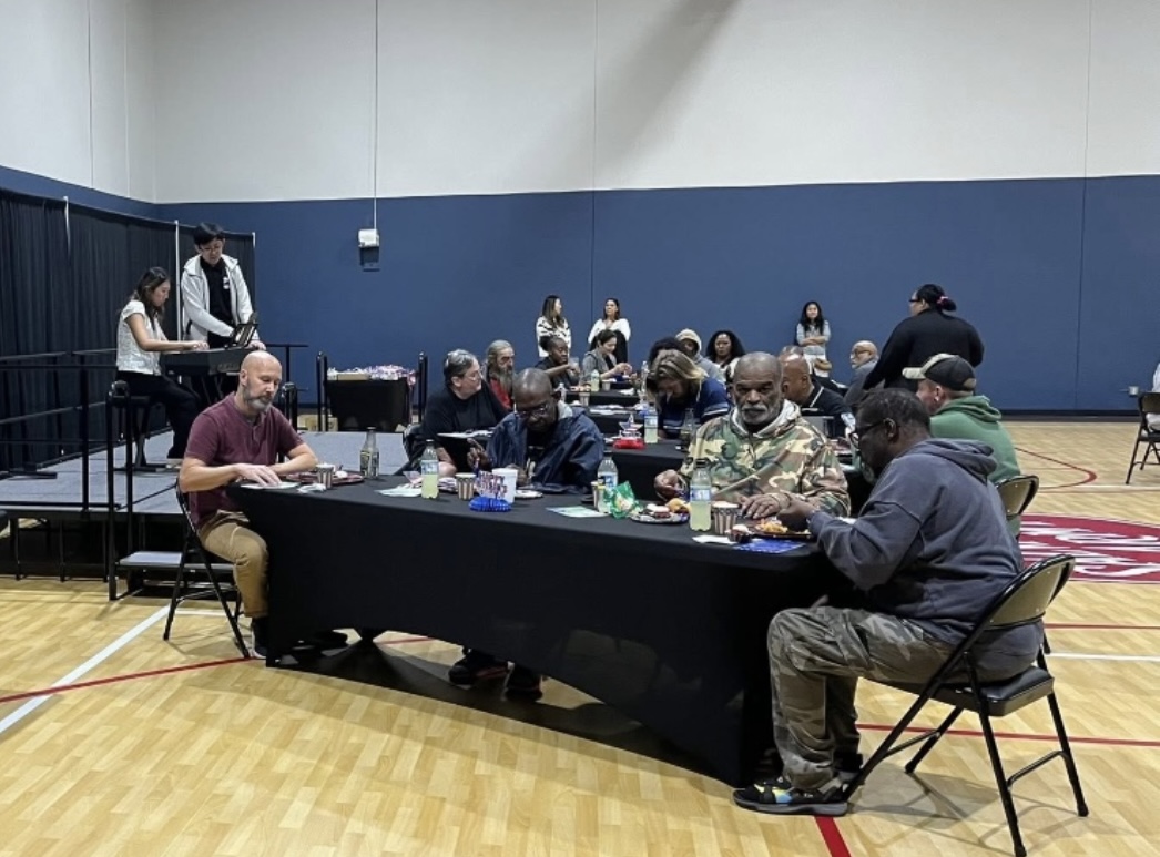 People seated at a long table in a gymnasium, with others standing and sitting in the background.