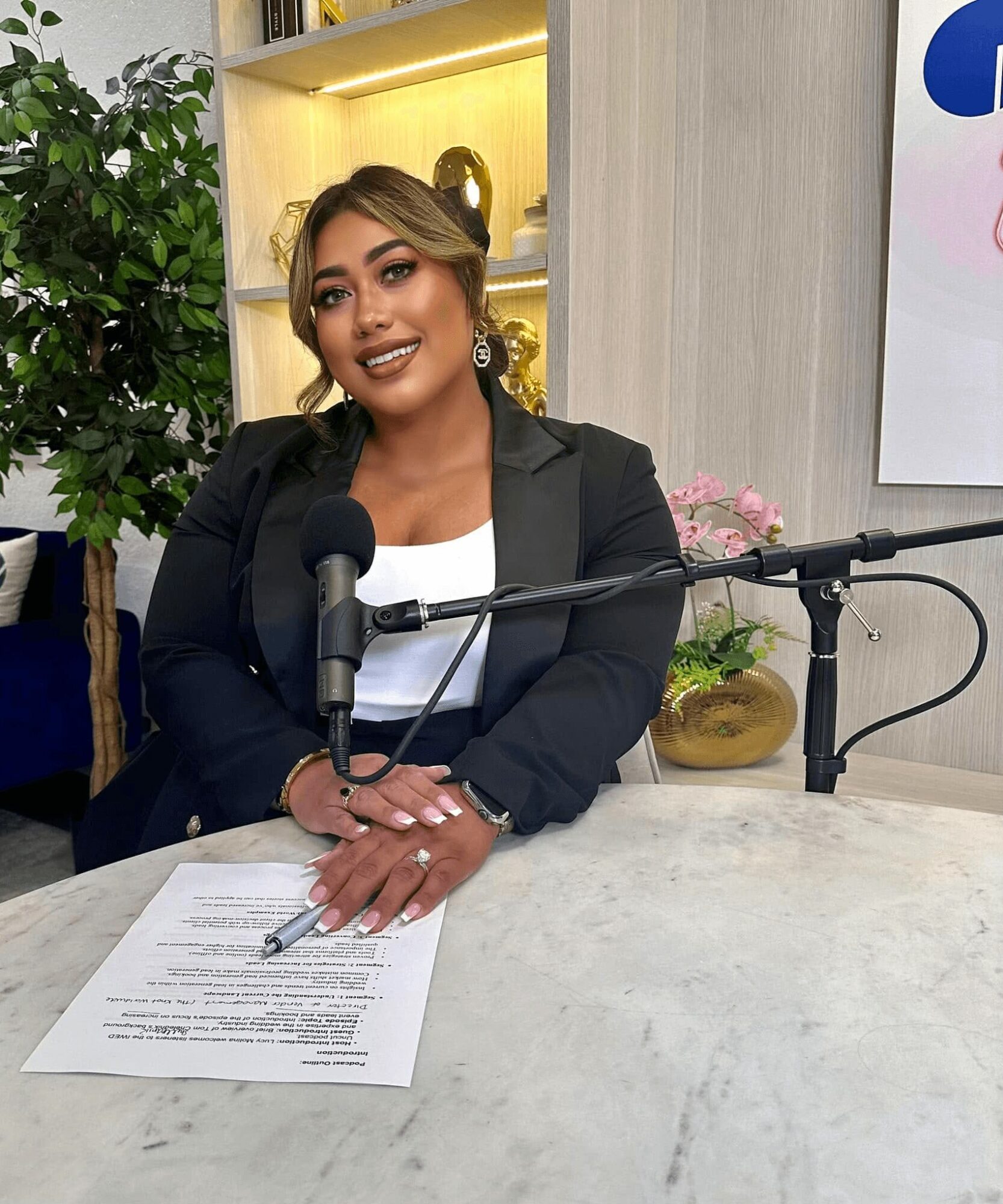 Woman with shoulder-length hair, wearing a black blazer and white top, sitting at a table with a microphone and papers.