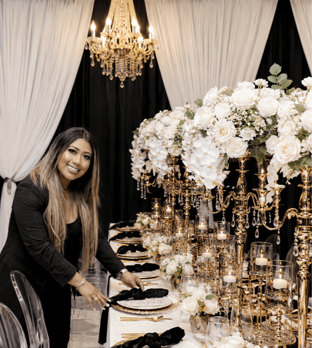 Woman smiling at a decorated table with white flowers and gold candelabras, chandelier overhead, curtains in background.