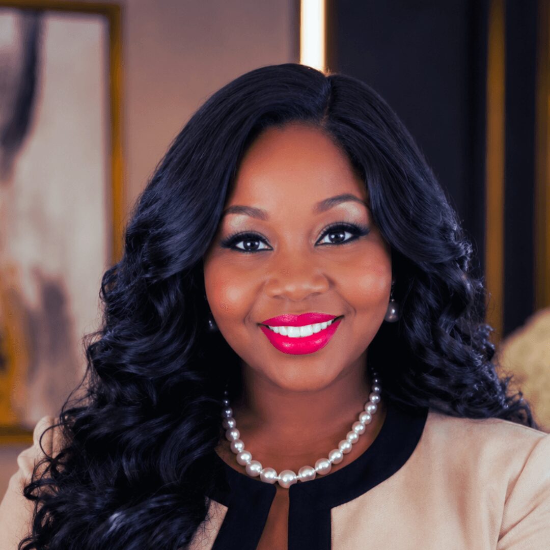 Portrait of a woman with long, curly black hair, wearing a pearl necklace and a beige top, smiling brightly.