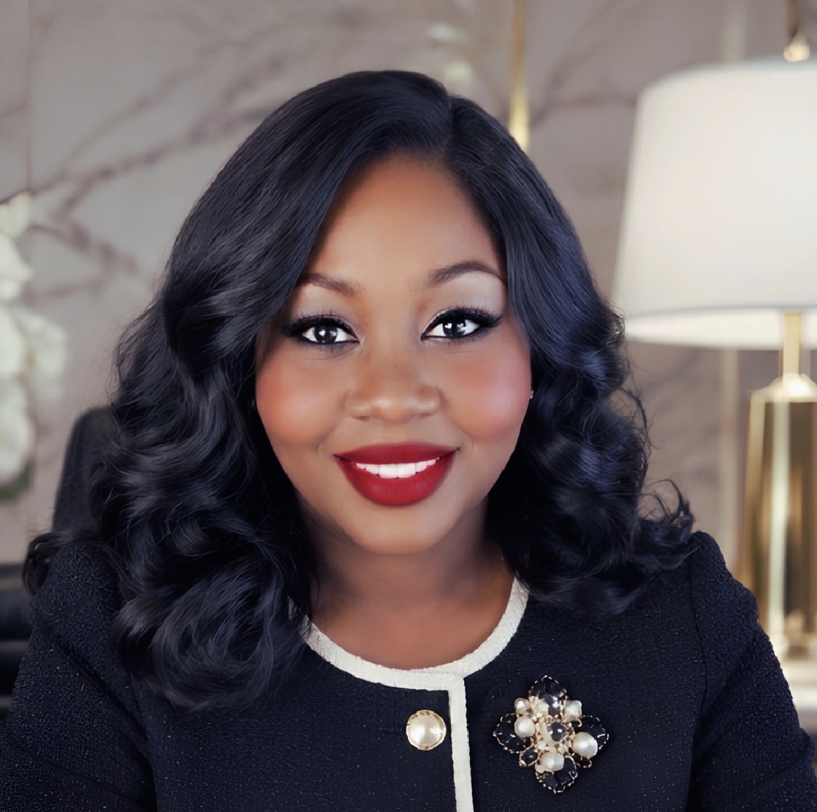 Woman with wavy black hair, red lipstick, wearing a black outfit with a decorative brooch, smiling indoors.